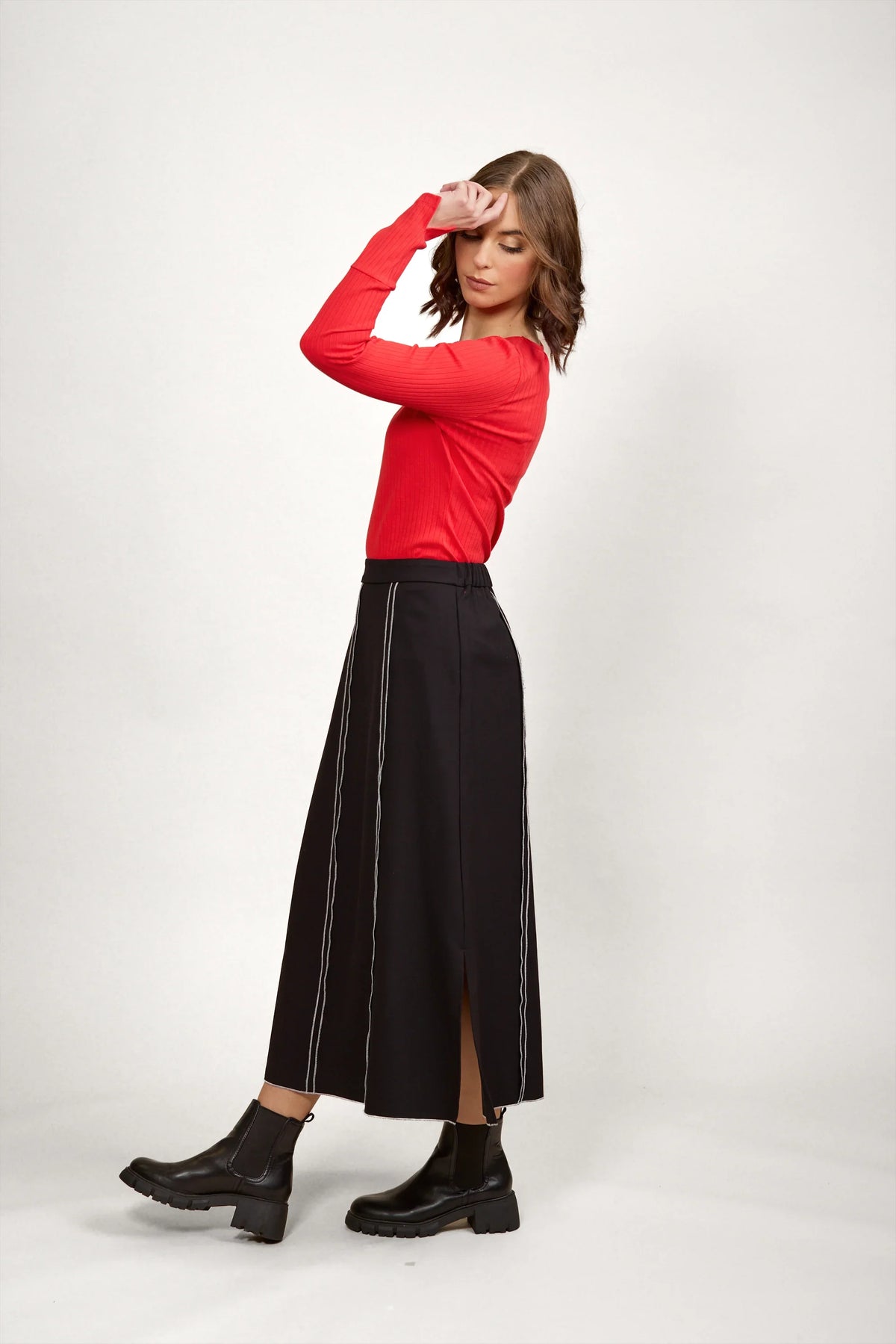 Woman in black midi skirt with white stitching, red long sleeve top, and black ankle boots