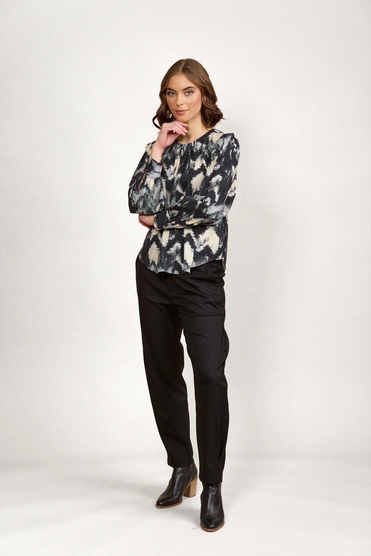 Woman wearing abstract print long sleeve blouse with black pants and ankle boots, studio background