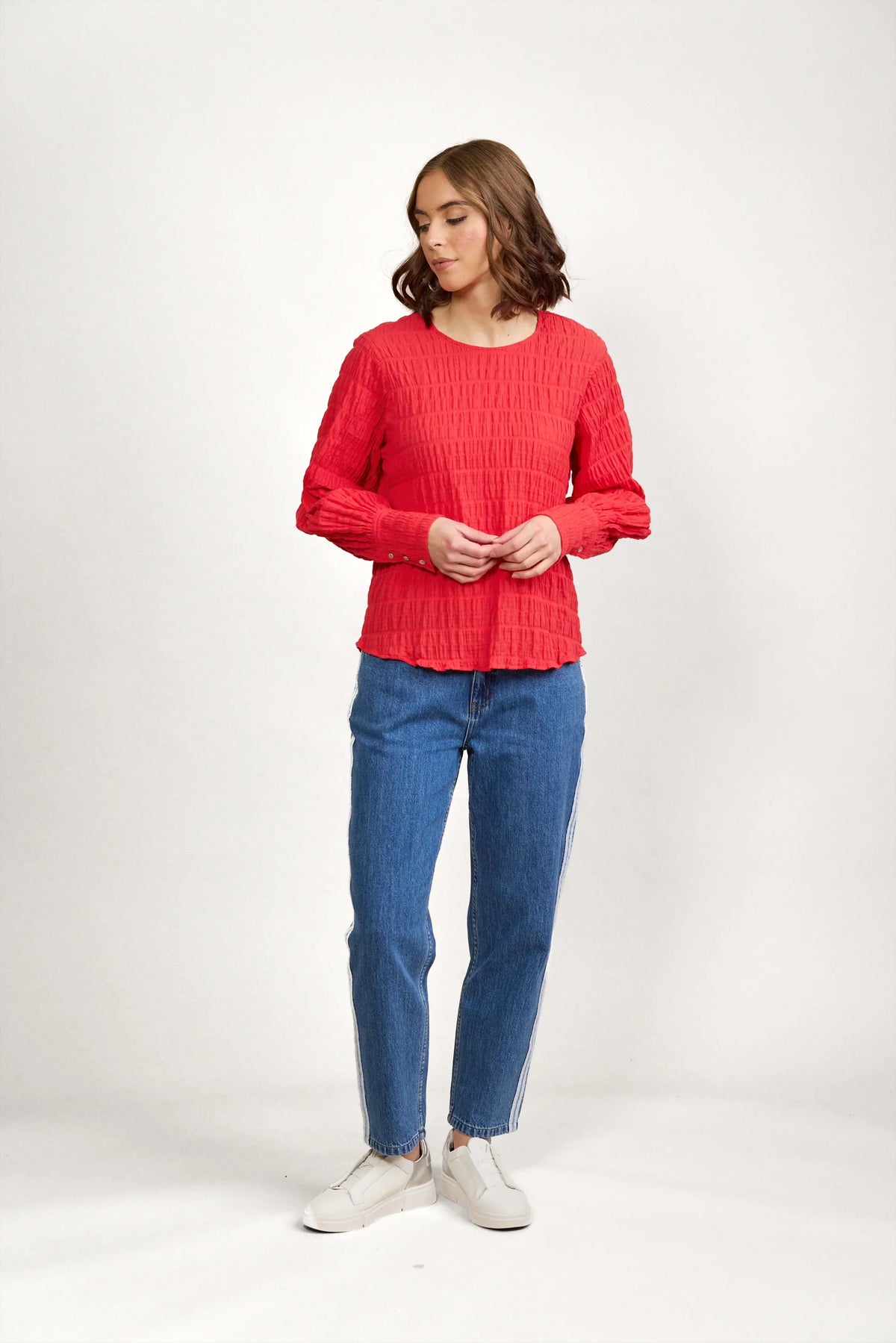 Woman wearing textured red long sleeve top, blue jeans, and white sneakers against plain background