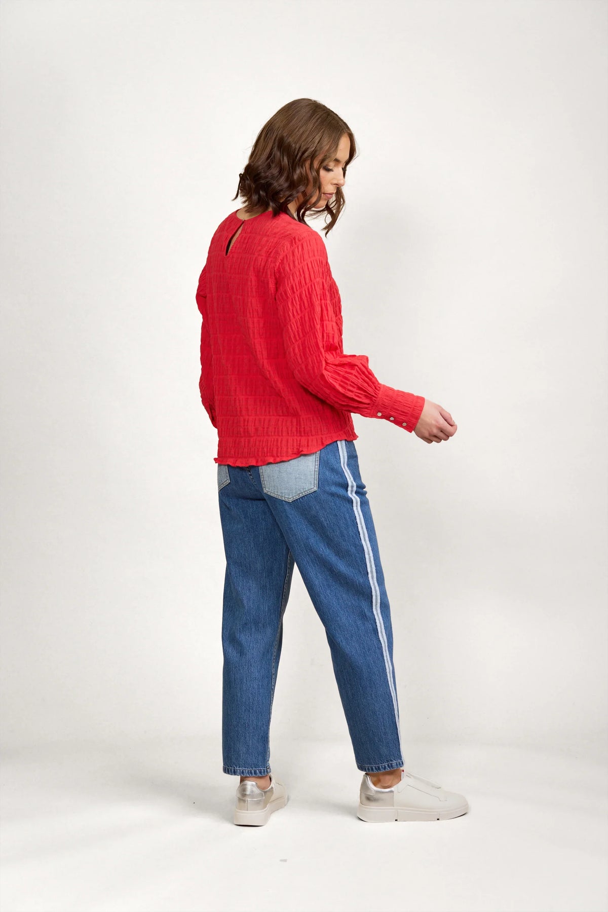 Woman in textured red long sleeve top, blue straight leg jeans with side stripe, and white sneakers