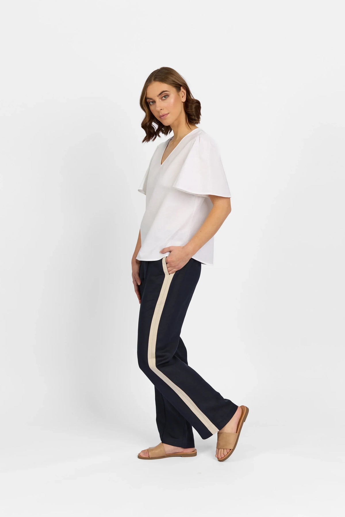 Woman wearing white flutter sleeve top and navy pants with beige stripe on white background