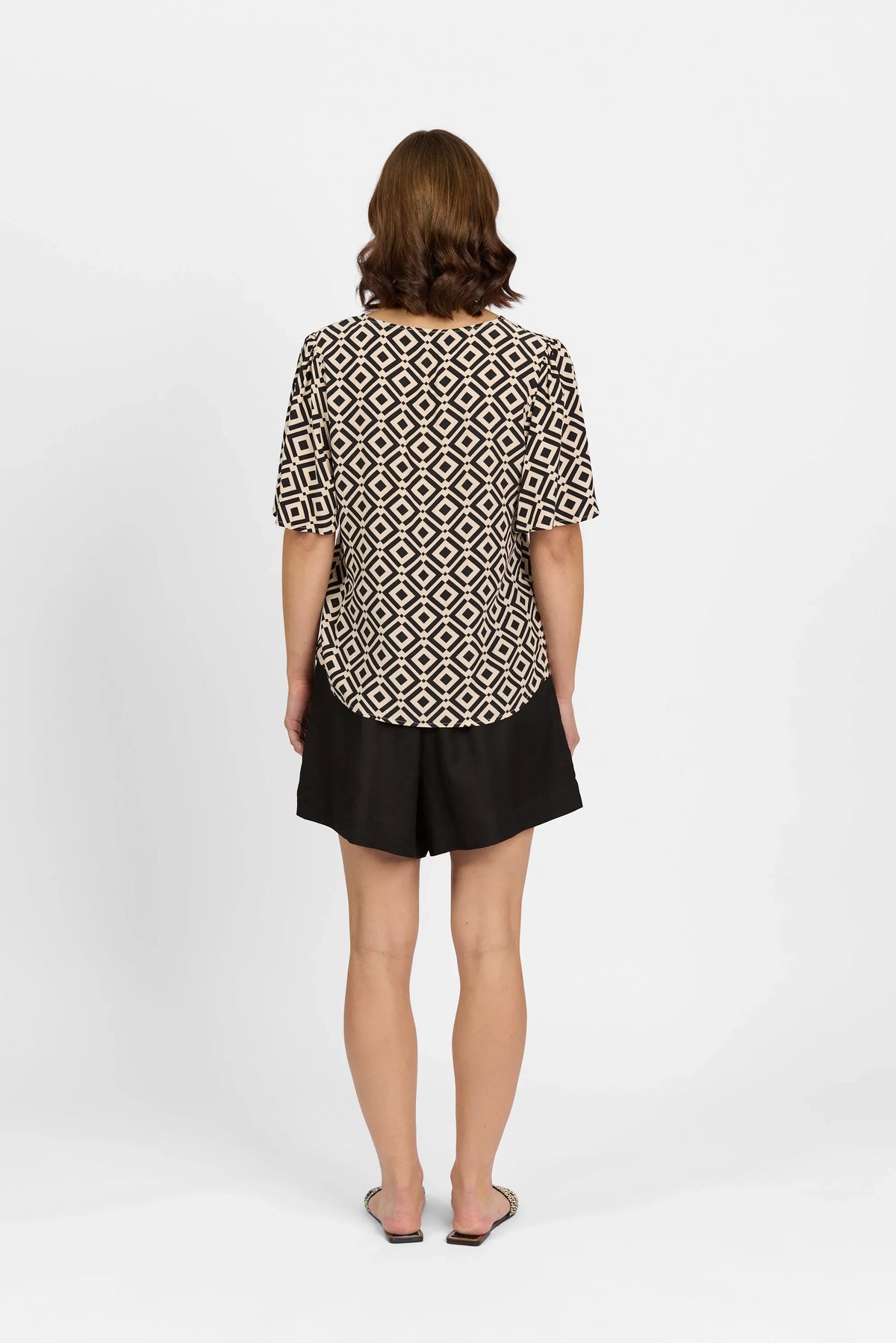 Woman wearing black and beige geometric print top with short sleeves and black shorts, back view