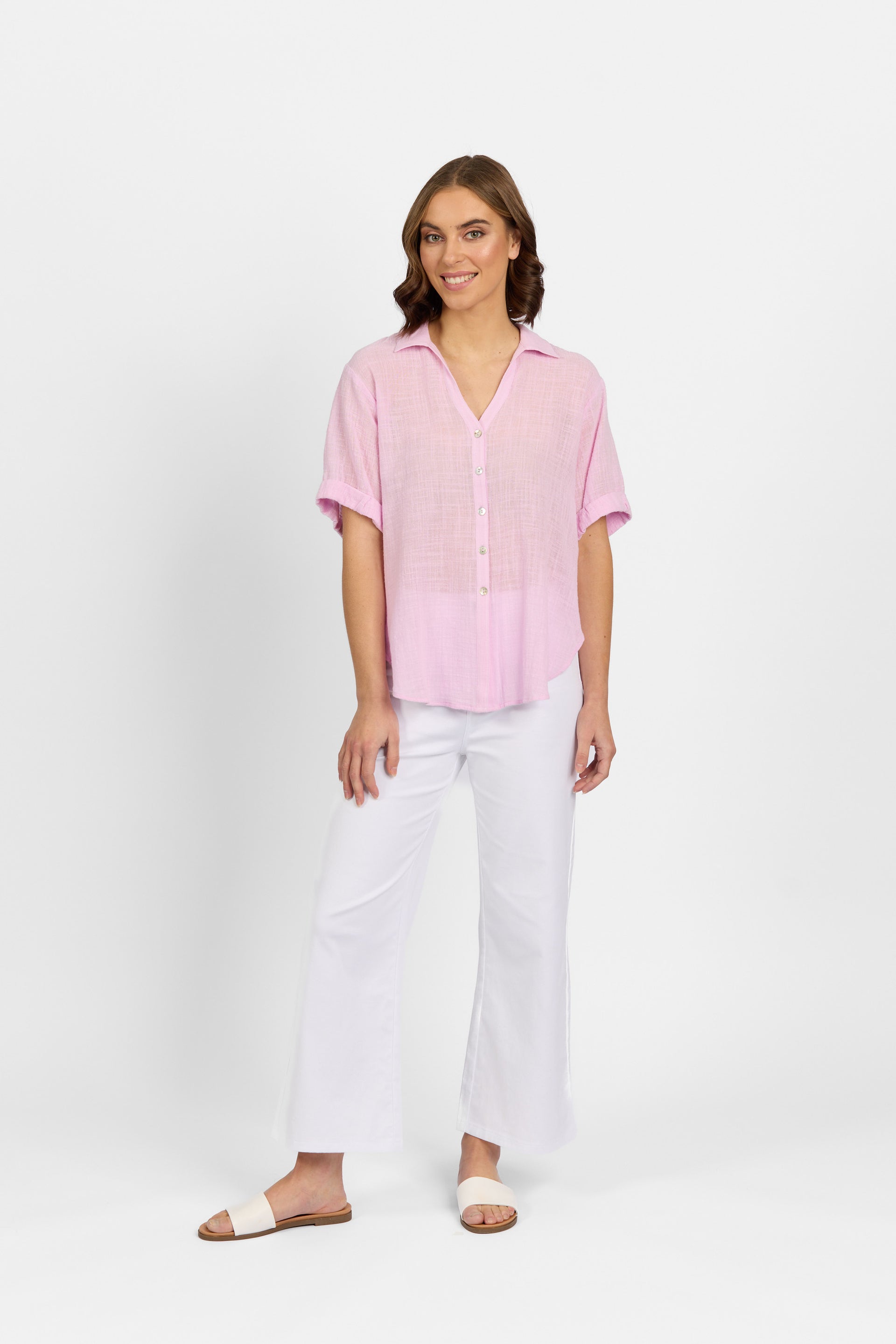 A woman with shoulder-length brown hair smiles gently against a white background, wearing Knewe Label's relaxed fit Infinity Shirt in Lilac paired with white wide-leg pants and slide sandals.