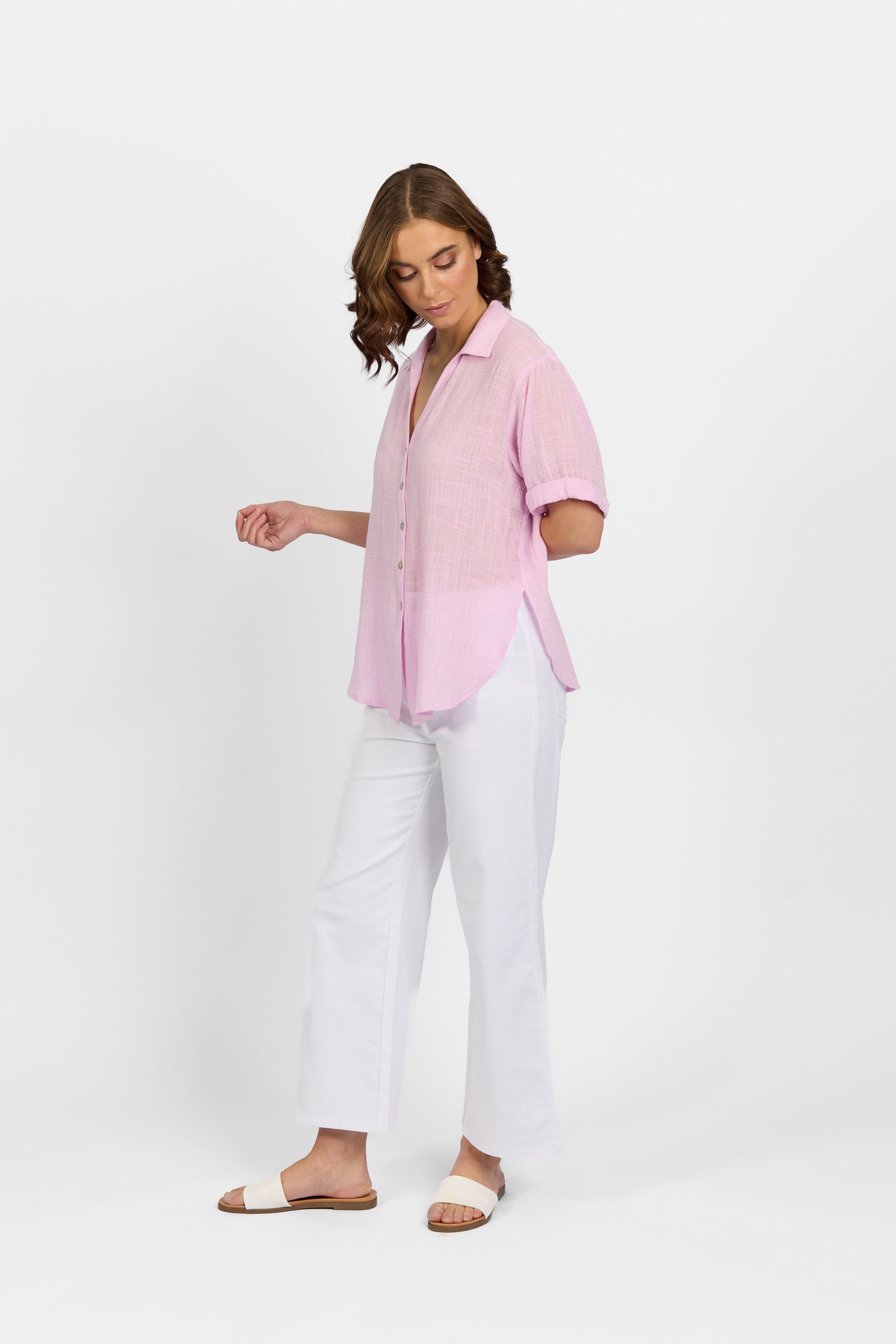A woman stands against a white background, wearing the Knewe Label Infinity Shirt in Lilac with a relaxed fit. She pairs it with white wide-leg pants and slide sandals, looking down and gently touching her shirt with one hand.