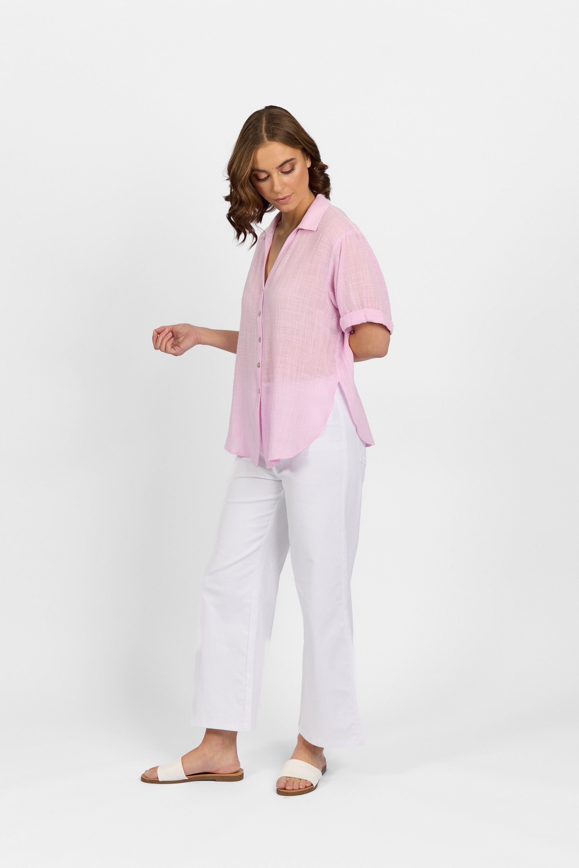 A woman stands against a white background, wearing the Knewe Label Infinity Shirt in Lilac with a relaxed fit. She pairs it with white wide-leg pants and slide sandals, looking down and gently touching her shirt with one hand.