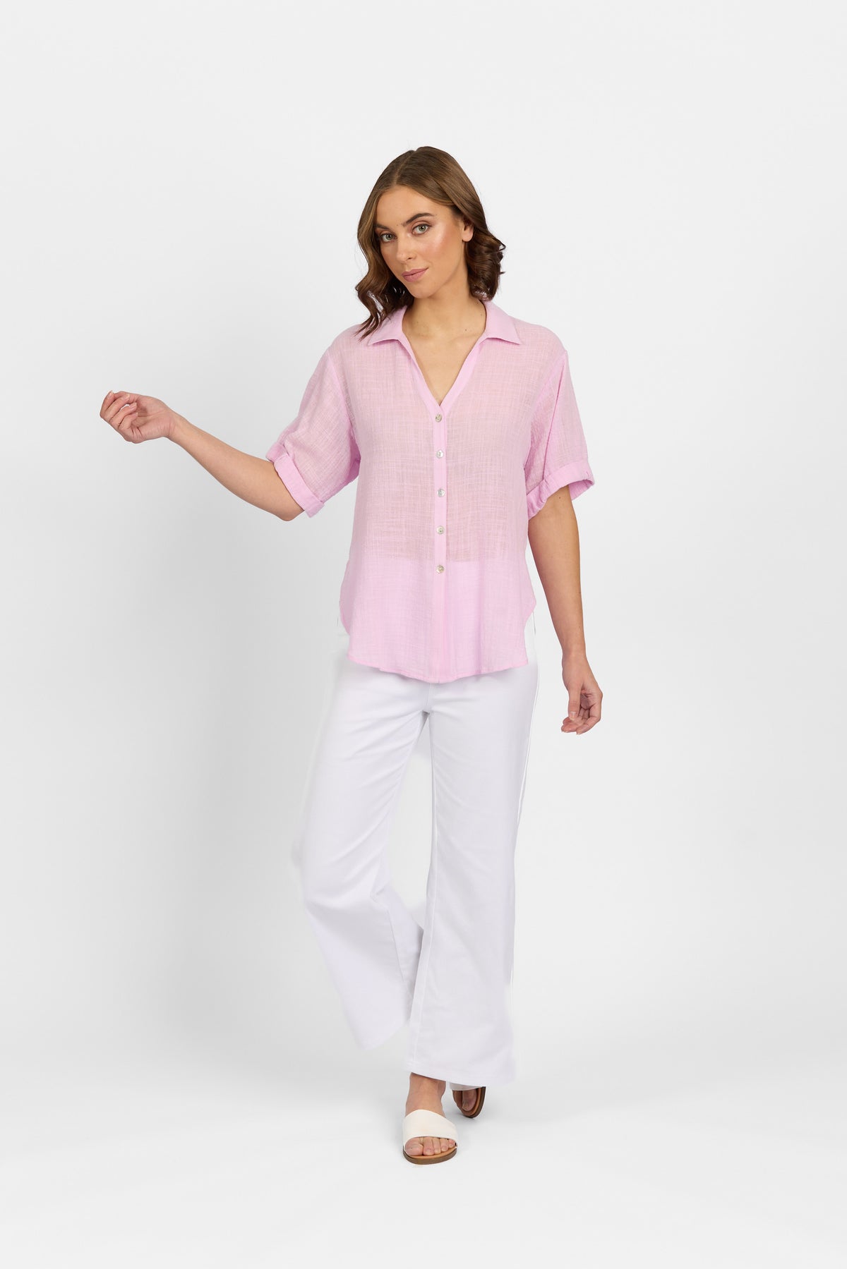A woman stands against a white background wearing the Knewe Label Infinity Shirt in lilac, crafted from lightweight cotton with a relaxed fit. She pairs it with white wide-leg pants and open-toe sandals, her wavy brown hair cascading as she looks left.
