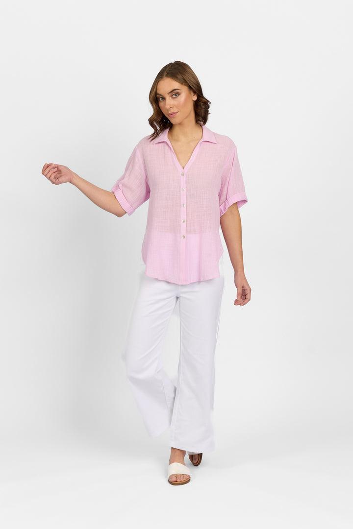 A woman stands against a white background wearing the Knewe Label Infinity Shirt in lilac, crafted from lightweight cotton with a relaxed fit. She pairs it with white wide-leg pants and open-toe sandals, her wavy brown hair cascading as she looks left.