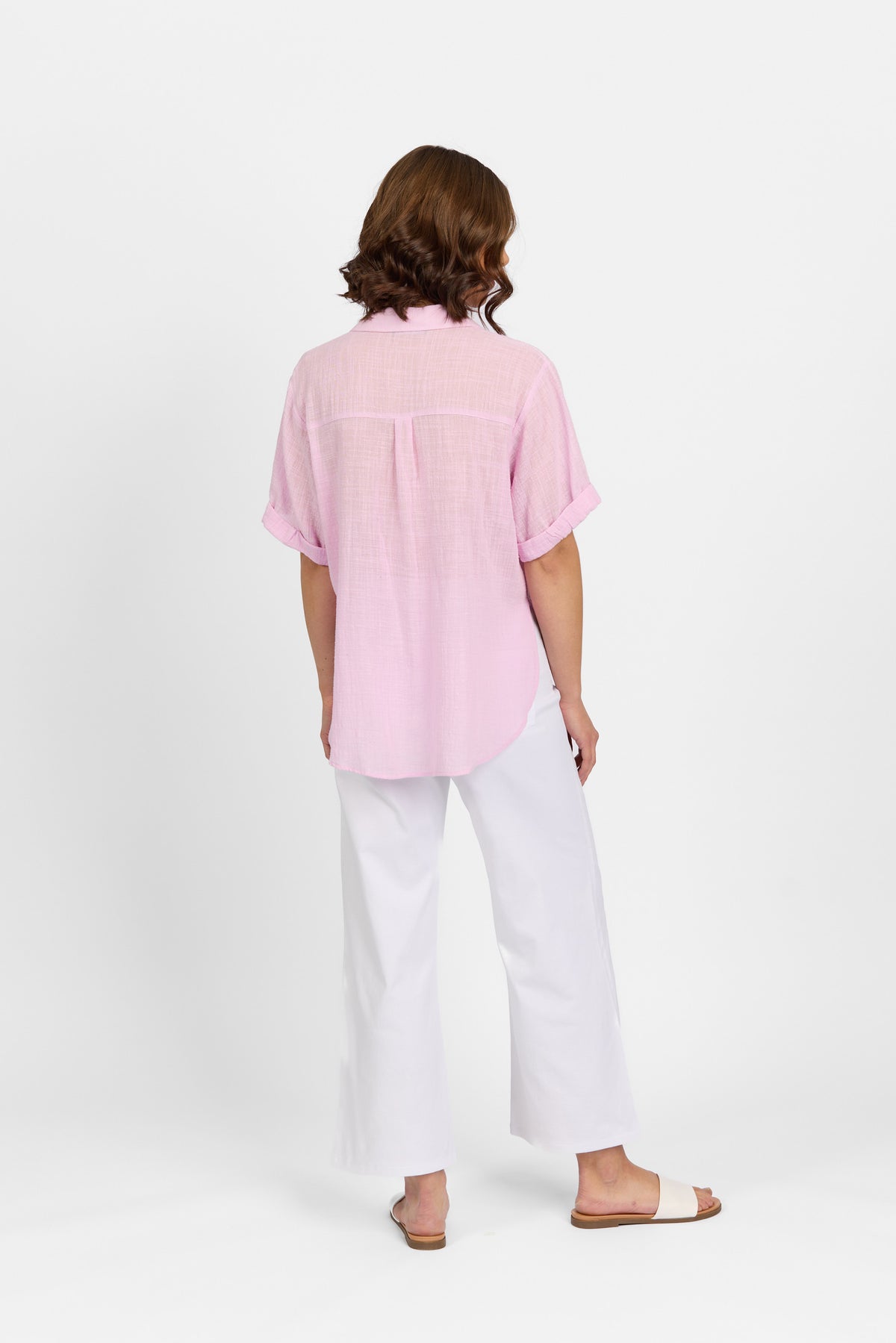 A person stands facing away, wearing a Knewe Label Infinity Shirt in Lilac (relaxed fit), loose white pants, and beige slide sandals, set against a plain white background.