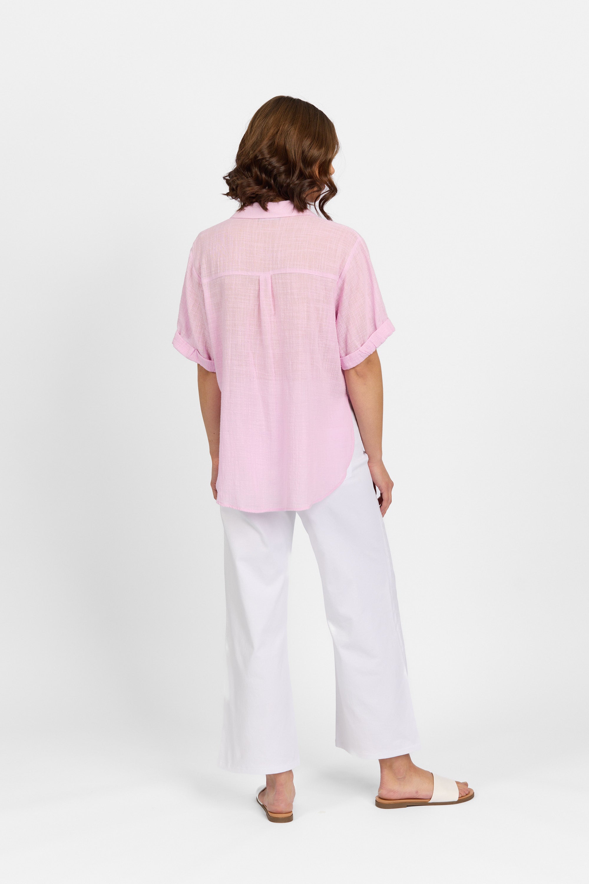 A person stands facing away, wearing a Knewe Label Infinity Shirt in Lilac (relaxed fit), loose white pants, and beige slide sandals, set against a plain white background.