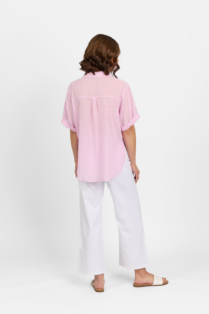 A person stands facing away, wearing a Knewe Label Infinity Shirt in Lilac (relaxed fit), loose white pants, and beige slide sandals, set against a plain white background.