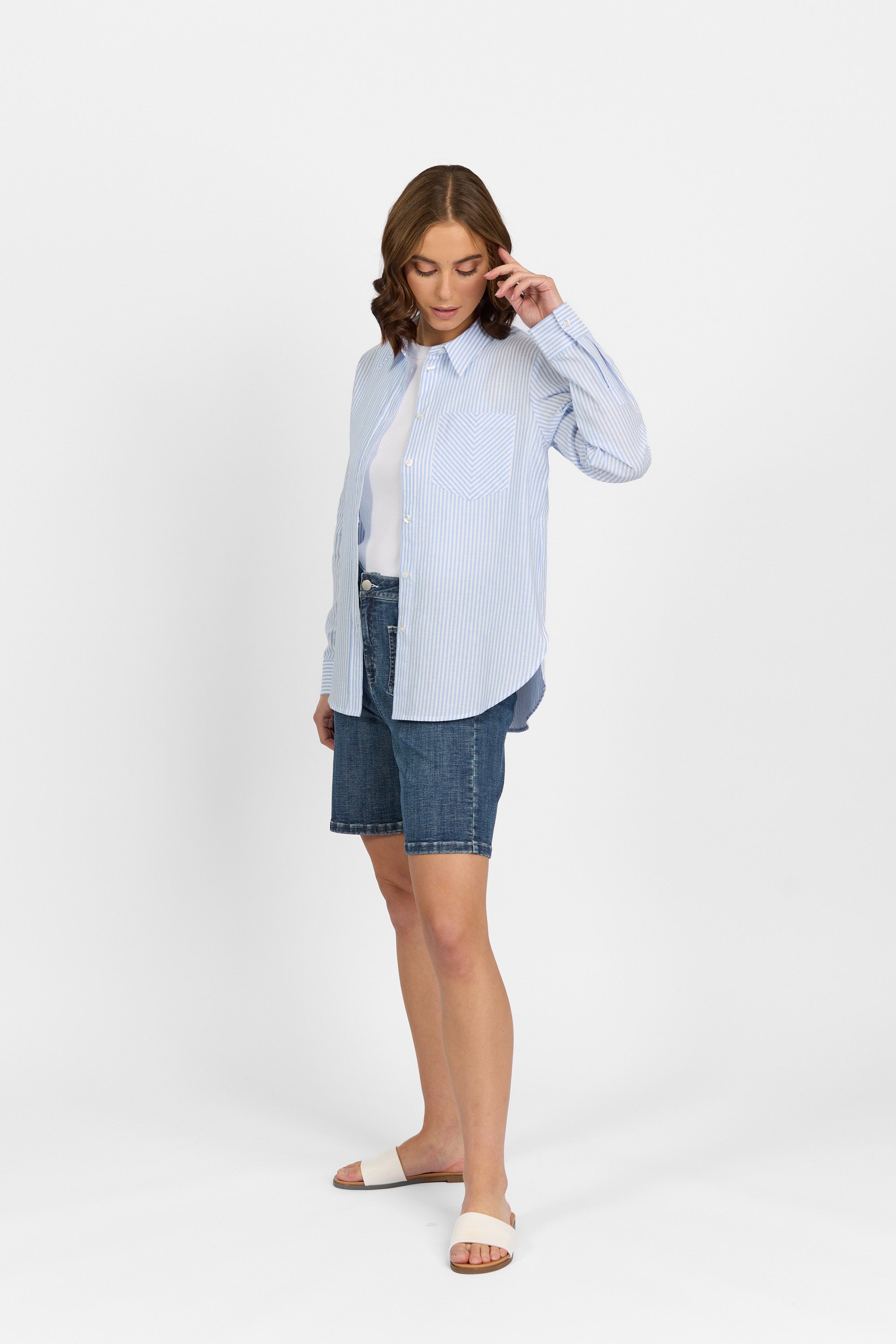 A woman stands against a white background, wearing the Knewe Label Tide Shirt in Blue Stripe over a white top, paired with denim knee-length shorts and white slide sandals. She looks down, gently touching her hair with one hand.