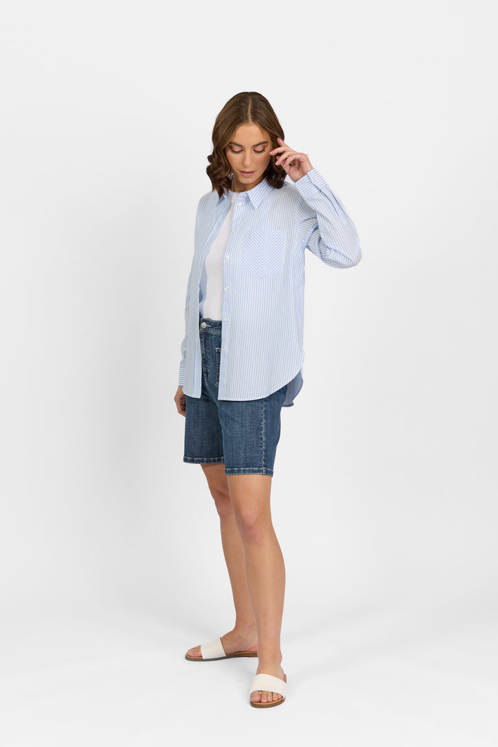 A woman stands against a white background, wearing the Knewe Label Tide Shirt in Blue Stripe over a white top, paired with denim knee-length shorts and white slide sandals. She looks down, gently touching her hair with one hand.