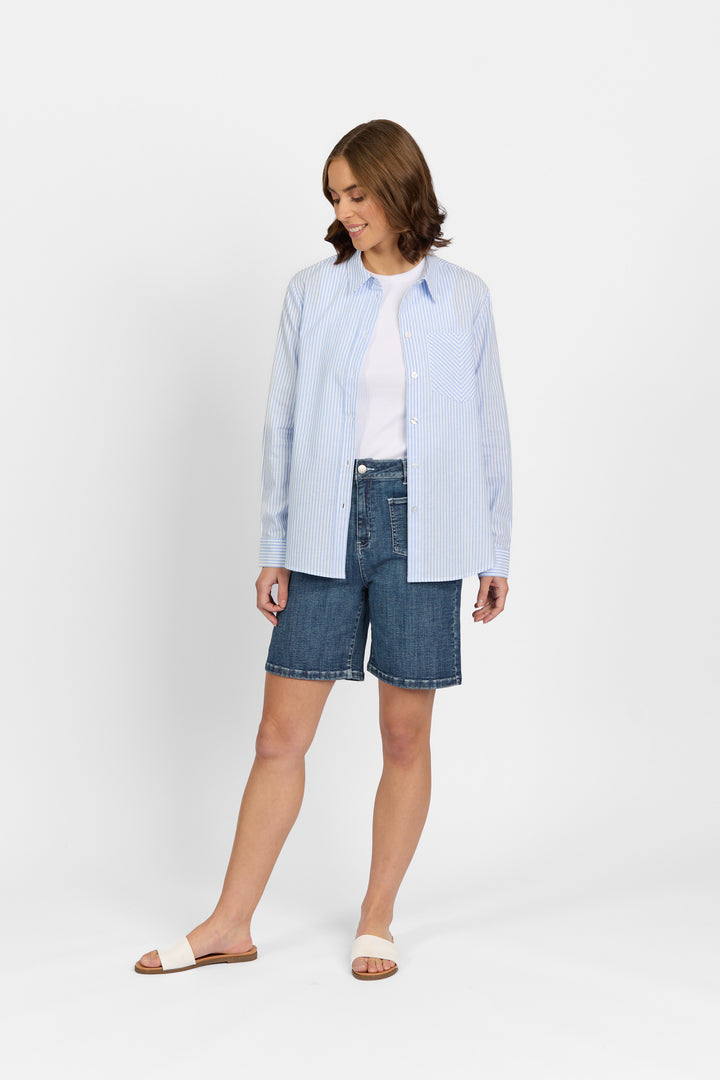 A woman smiles in Knewe Label's Tide Shirt - Blue Stripe, layered over a white tee, with denim knee-length shorts and white slide sandals, standing against a plain white background.
