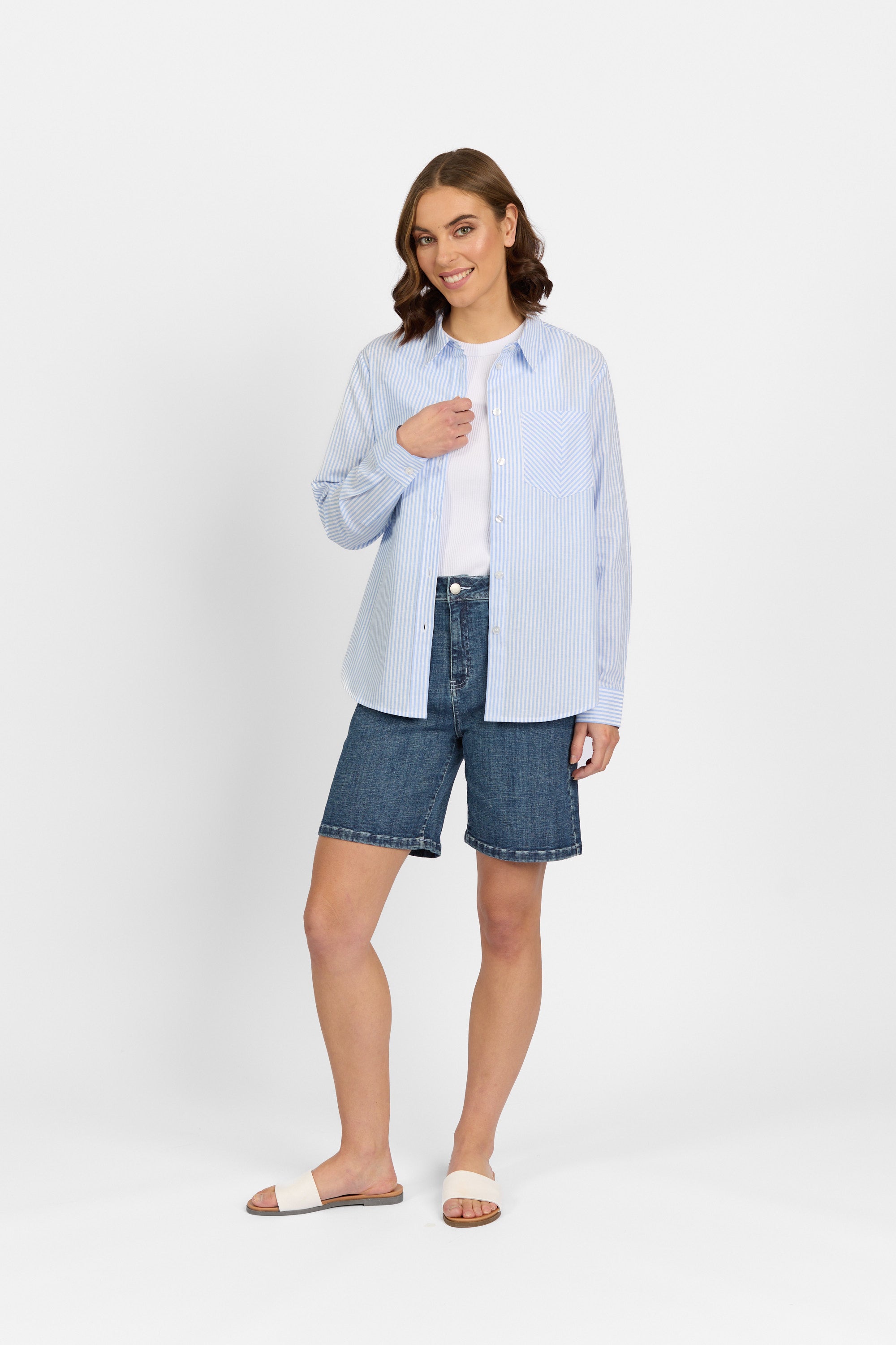 A woman with short brown hair smiles in a Knewe Label Tide Shirt - Blue Stripe over a white top, blue denim shorts, and white slide sandals, standing against a plain white background.