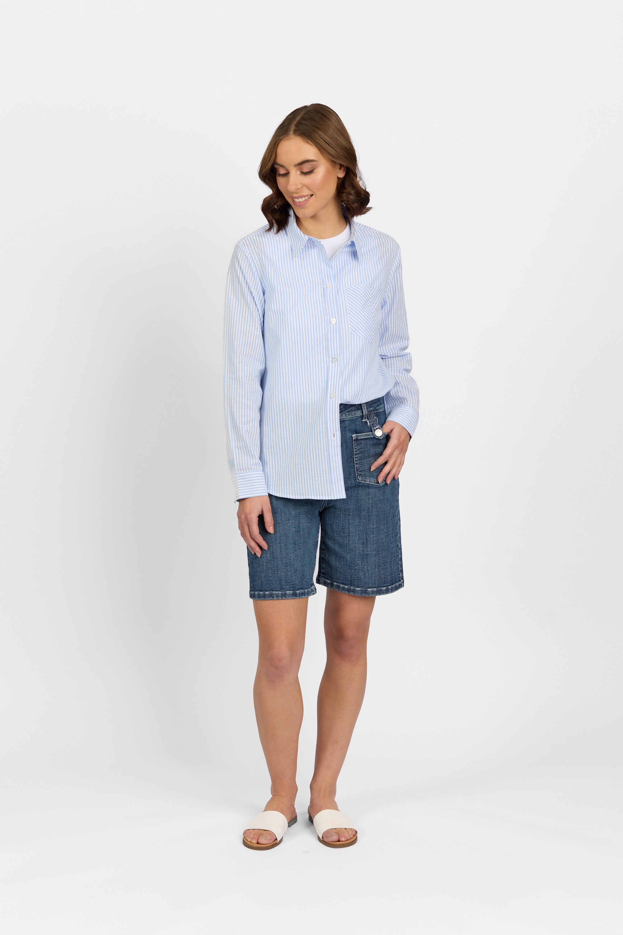 A woman stands against a white background, wearing the Knewe Label Tide Shirt - Blue Stripe with denim knee-length shorts and white slide sandals, looking down with a gentle smile.