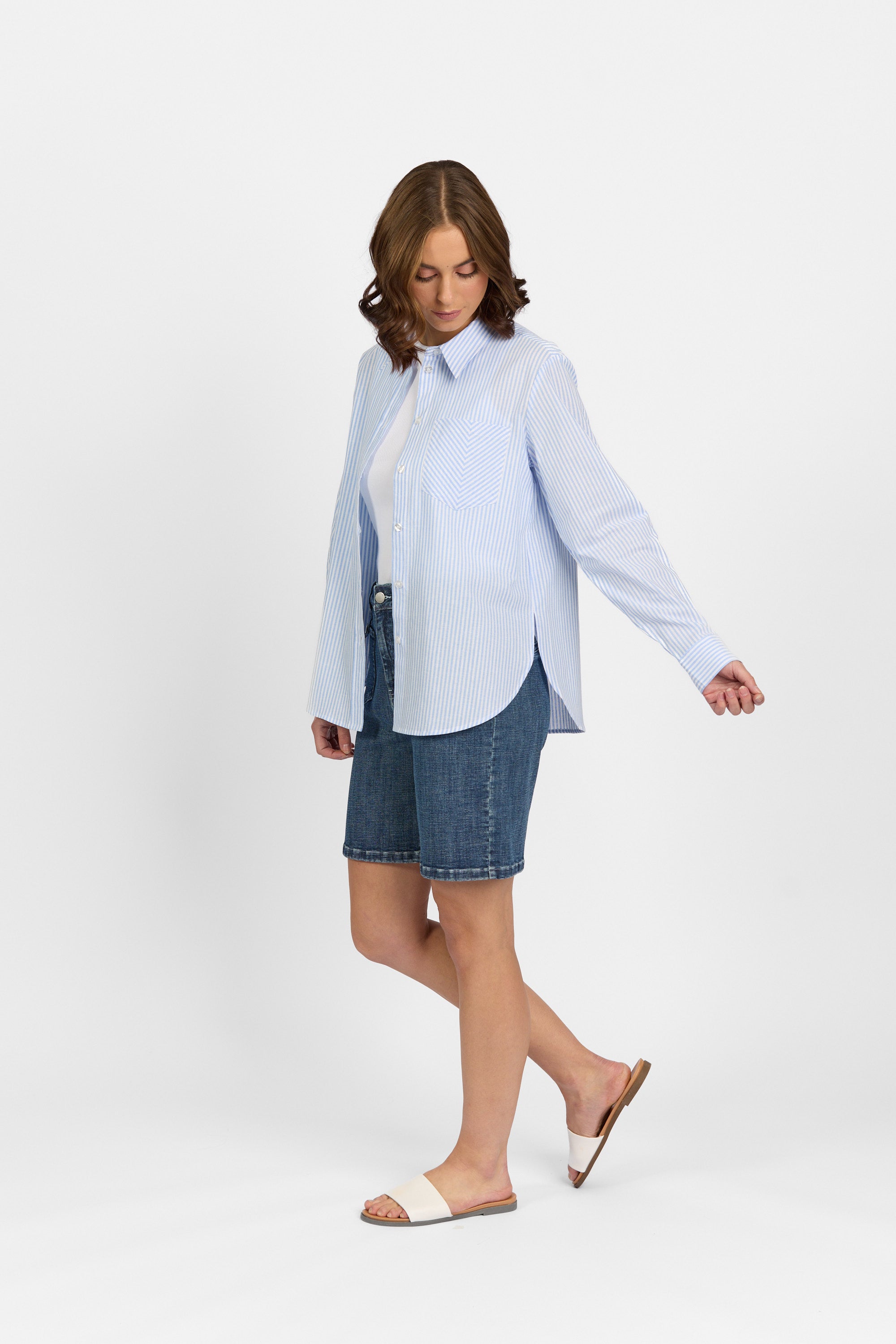 A woman wearing the Knewe Label Tide Shirt - Blue Stripe, a lightweight cotton top with blue stripes, paired with denim knee-length shorts and white slide sandals stands against a plain white background, looking down with one arm slightly out.