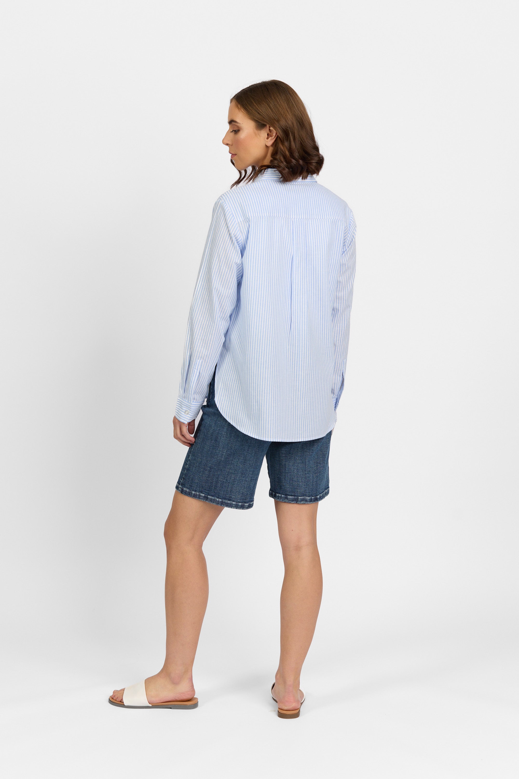 A person faces away, wearing the Knewe Label Tide Shirt - Blue Stripe with denim shorts and beige slide sandals, set against a plain white background.
