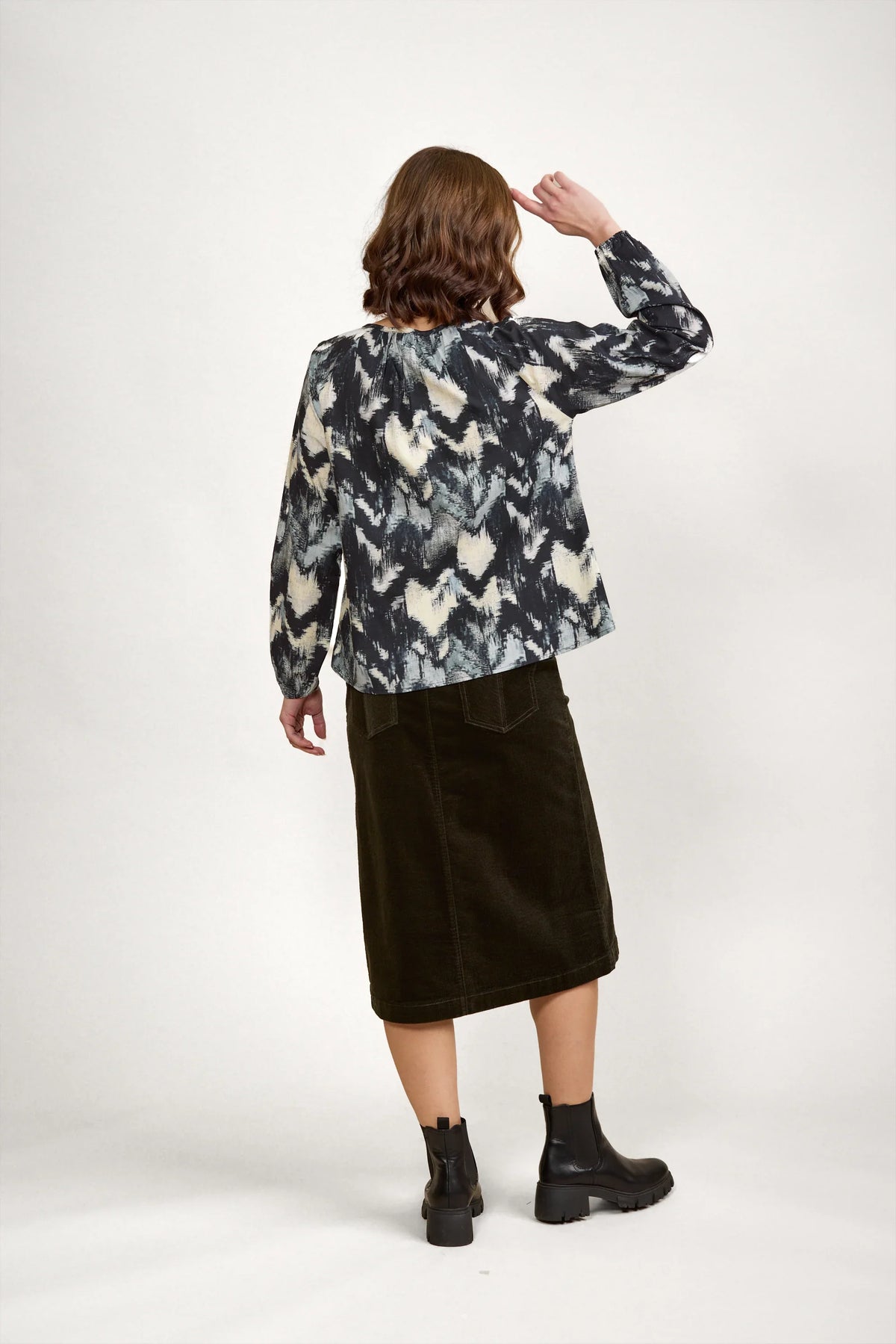 Woman in patterned long sleeve blouse, black midi skirt, and ankle boots, back view