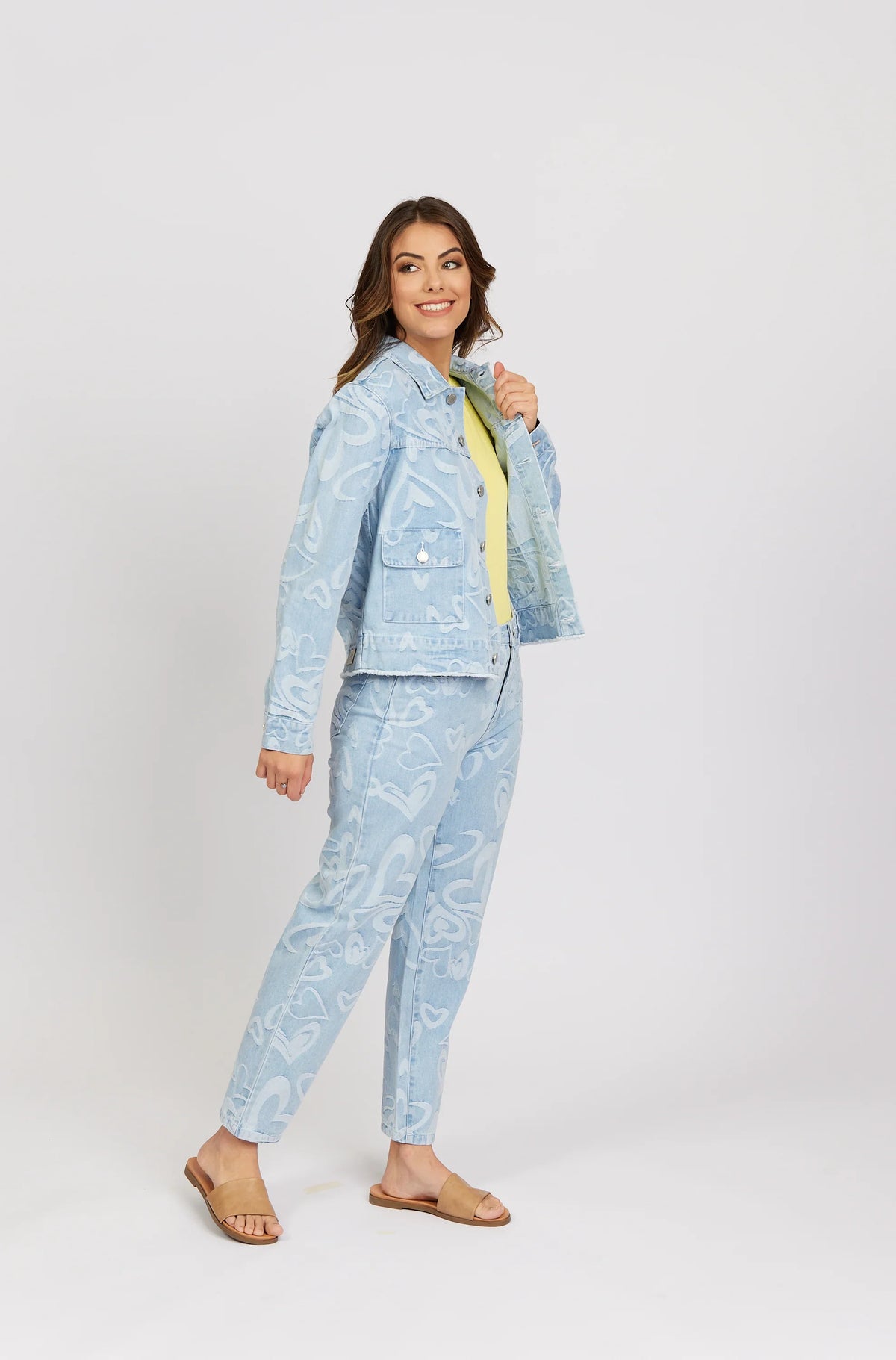 Woman modeling light blue denim jacket and jeans with heart patterns, yellow top, and sandals