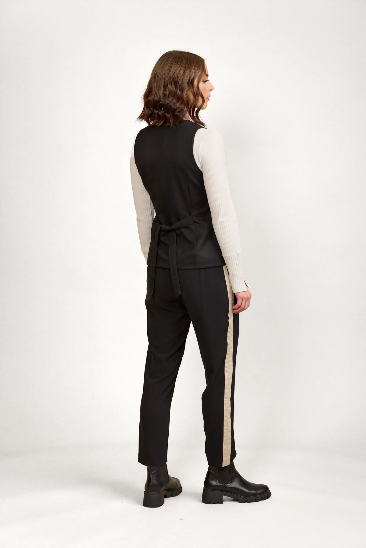 Woman in black tailored vest and black pants with beige side stripe, black boots, studio backdrop