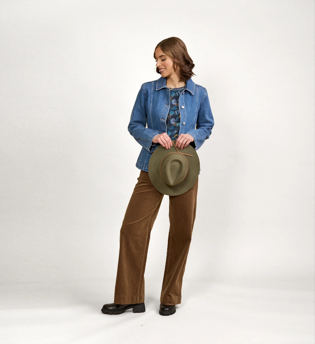 Woman modeling blue denim jacket with brown wide-leg pants, holding a green hat