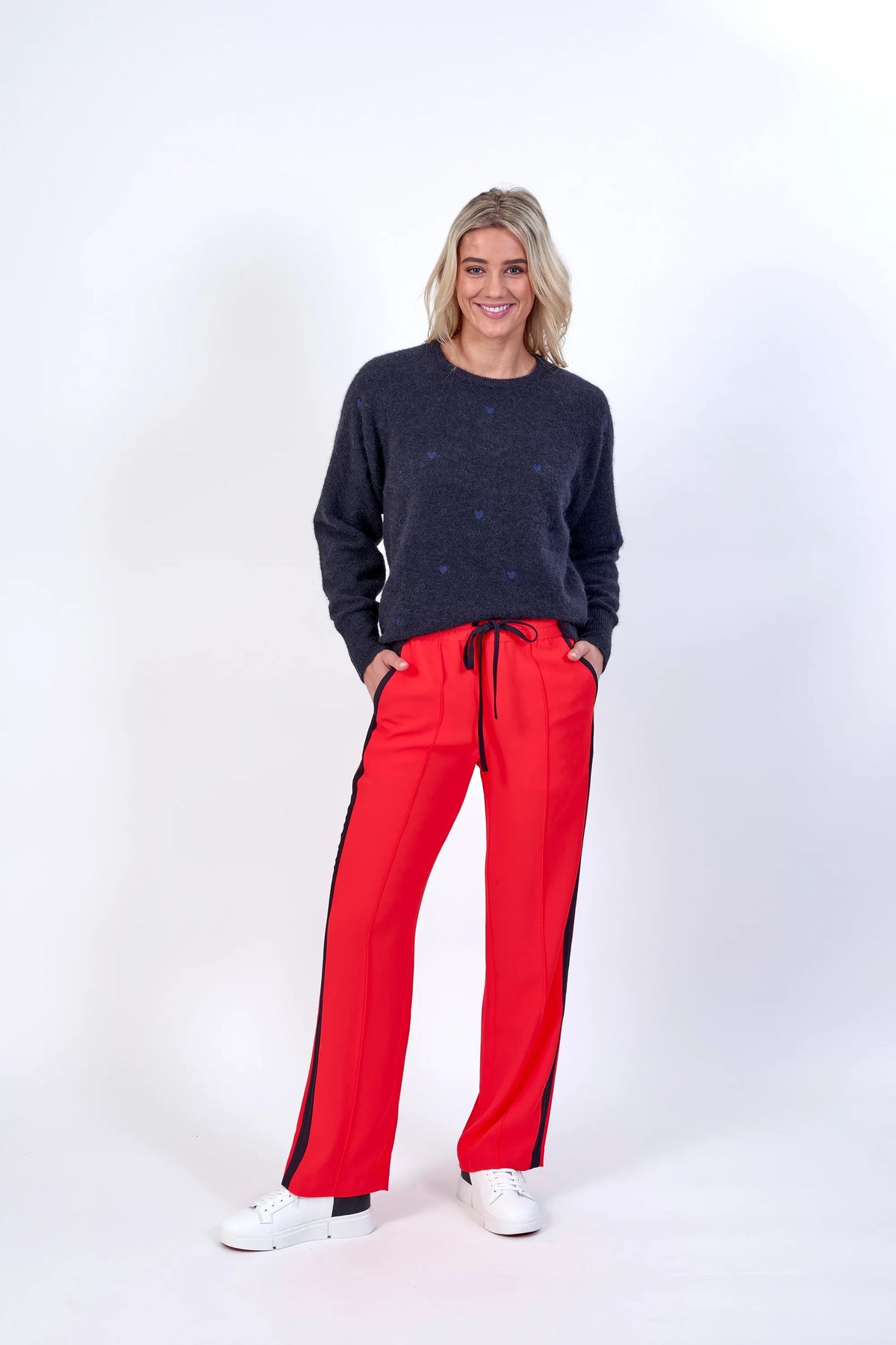 Woman wearing bright orange wide-leg pants with black side stripe and drawstring waist, paired with a navy sweater and white sneakers, studio background.