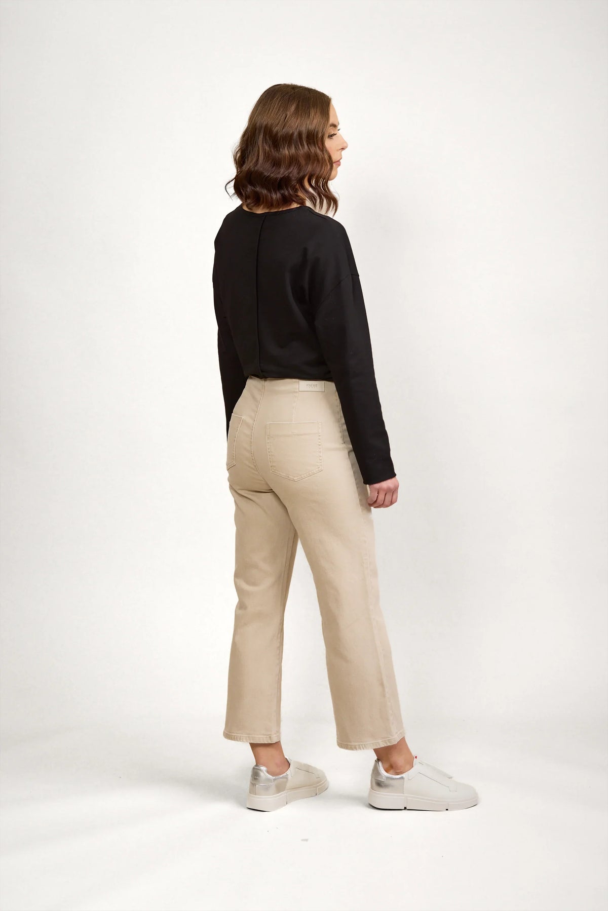 Woman wearing oat-colored denim jeans with straight leg, black long sleeve top, and white sneakers, standing against a neutral background