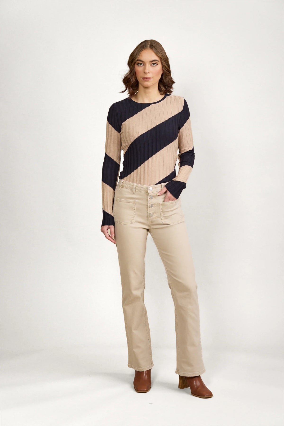 A woman with shoulder-length brown hair and a neutral expression stands against a white background, wearing Knewe Label's Potter Jean - Oat Denim bootcut pants, a beige and navy striped long-sleeve top, and brown ankle boots.