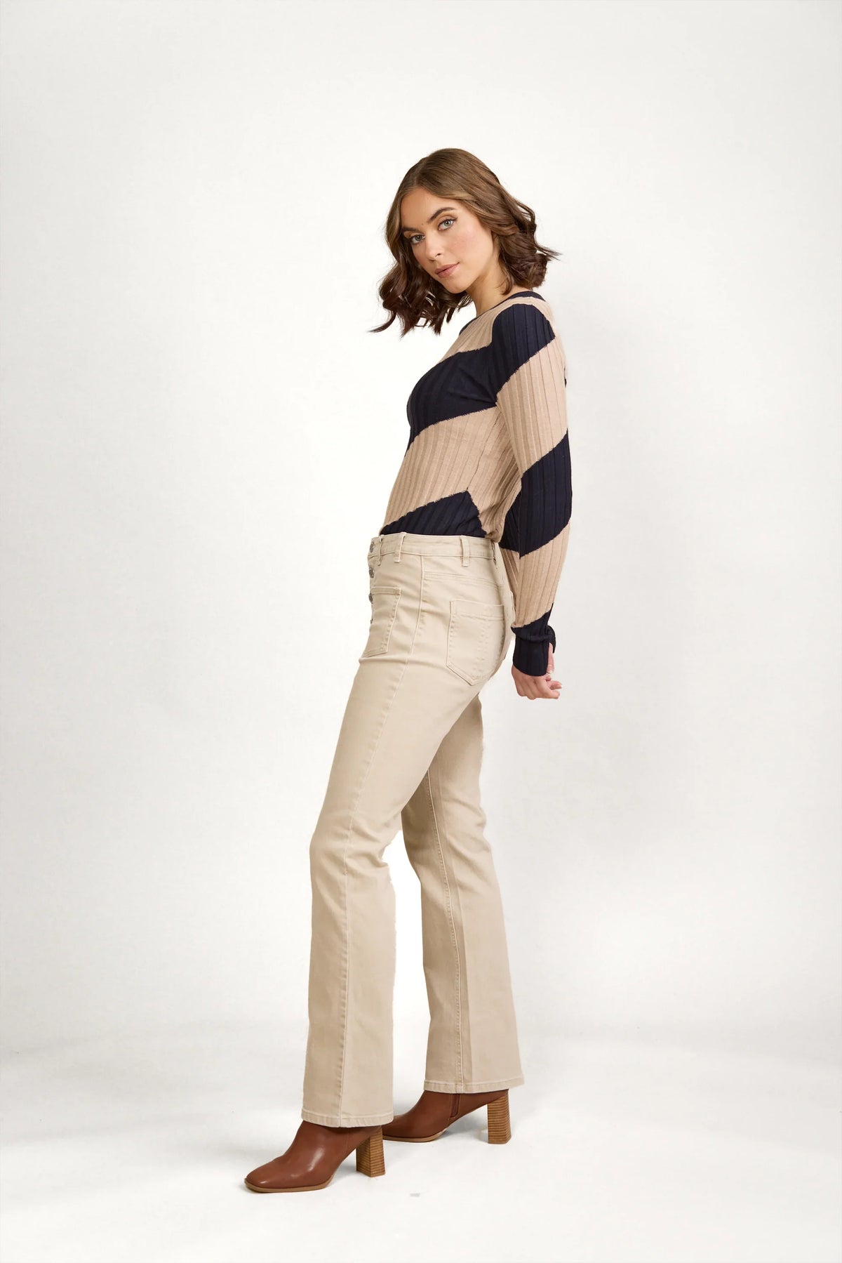 A woman with shoulder-length brown hair stands sideways, looking at the camera. She wears a tan and navy striped long-sleeve top, Knewe Label Potter Jean in Oat Denim, and brown heeled boots against a plain white background.