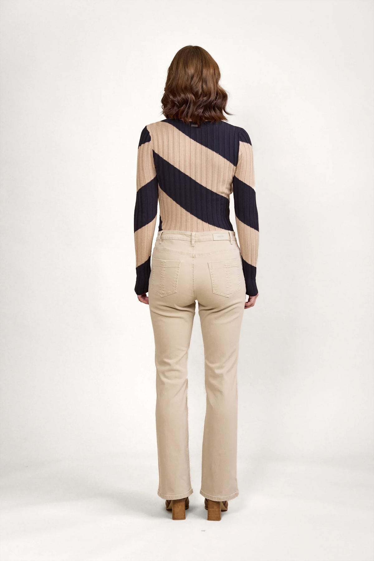 A woman stands facing away against a white background, wearing a long-sleeve diagonally striped black and beige sweater, Knewe Label's Potter Jean in Oat Denim, and heeled shoes.