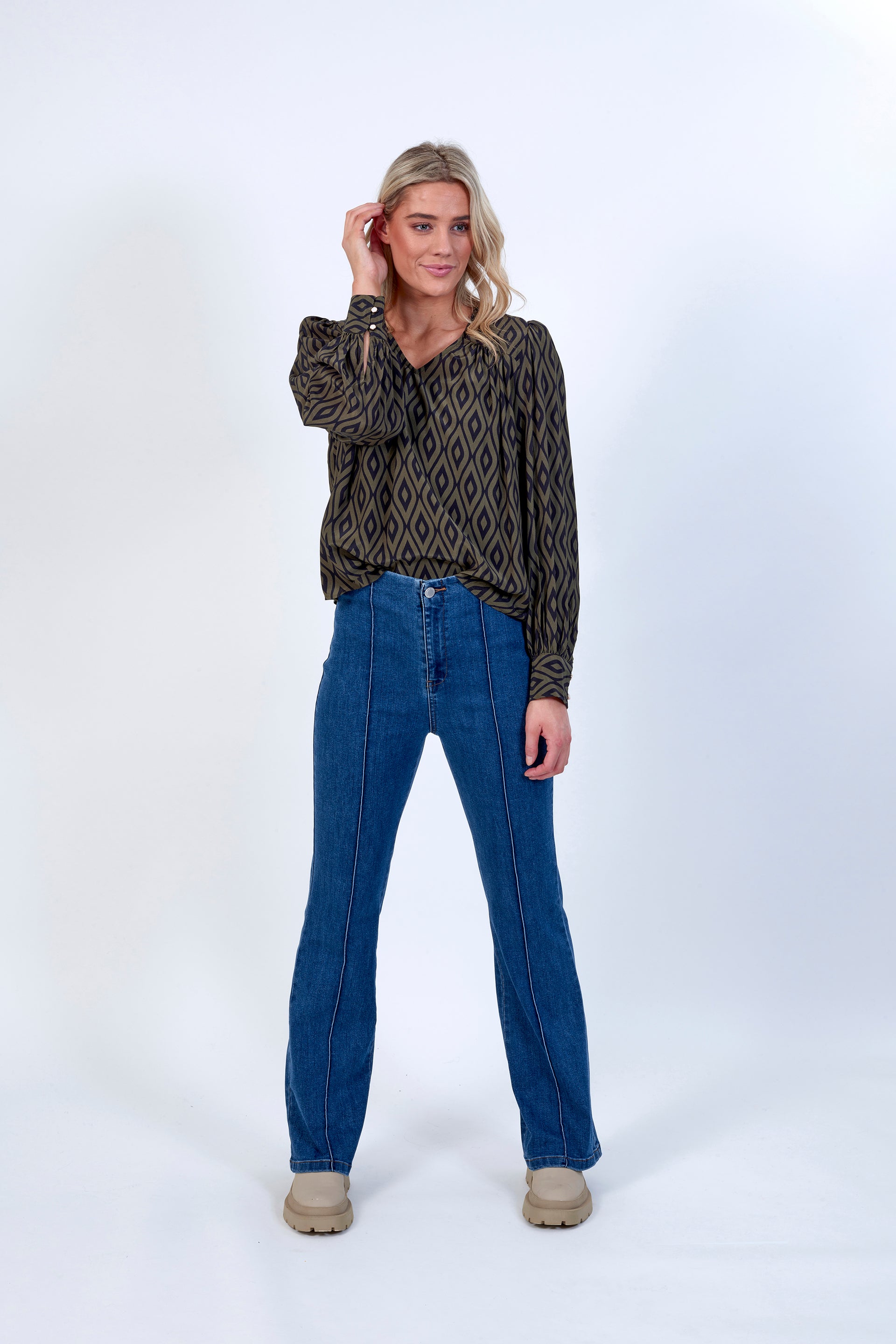 A woman with blonde hair smiles against a white background, wearing a patterned long-sleeve blouse, Knewe Label Haze Jean - Indigo high-waisted blue jeans with front seams in stretch fabric, and beige platform shoes.