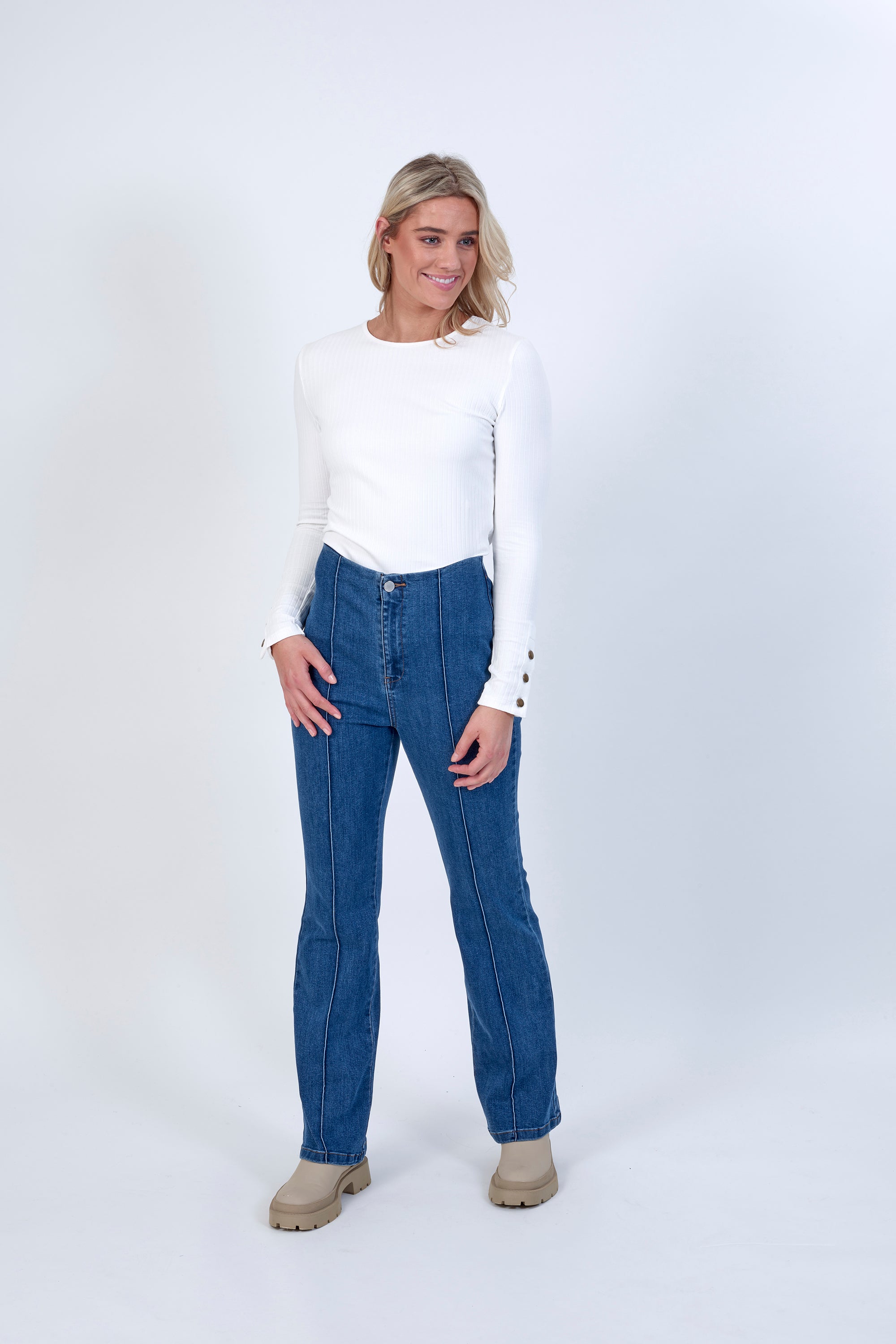 A woman with blonde hair smiles against a white background wearing a white long-sleeve top, Knewe Label Haze Jean - Indigo bootcut pants in blue stretch fabric, and beige ankle boots.