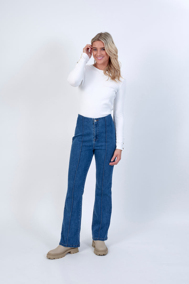 A woman with long blonde hair smiles against a white background, wearing a white long-sleeve top, Knewe Label’s Haze Jean – Indigo in stretch fabric, and beige boots. One hand touches her head.