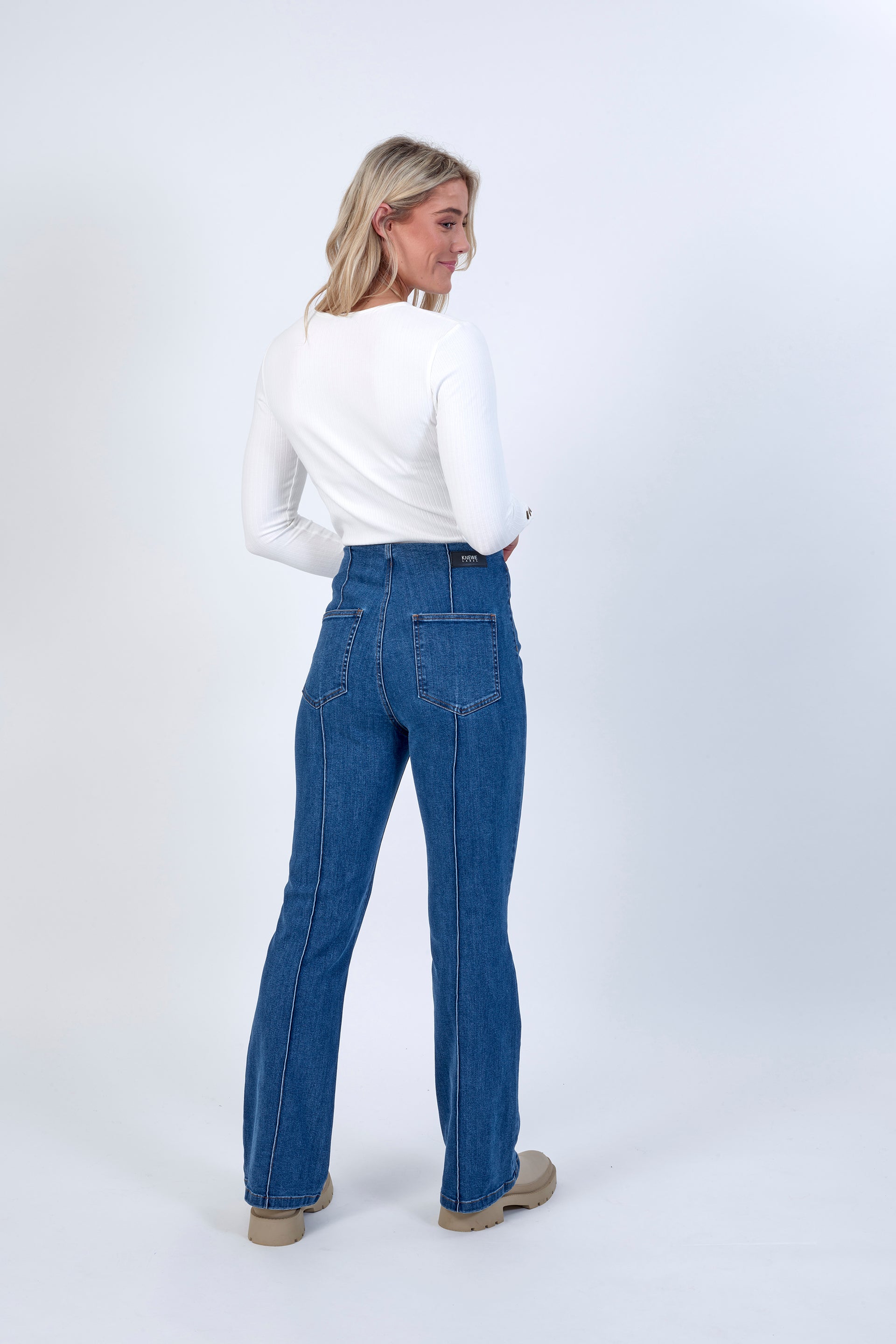 A woman with blonde hair faces away, glancing over her shoulder in a white long-sleeve top and Knewe Label’s Haze Jean - Indigo bootcut pants with beige shoes, set against a plain light background.
