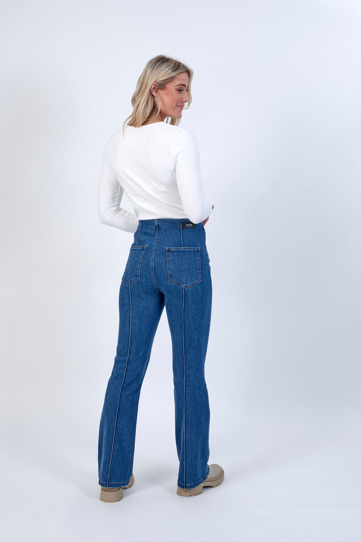 A woman with blonde hair faces away, glancing over her shoulder in a white long-sleeve top and Knewe Label’s Haze Jean - Indigo bootcut pants with beige shoes, set against a plain light background.