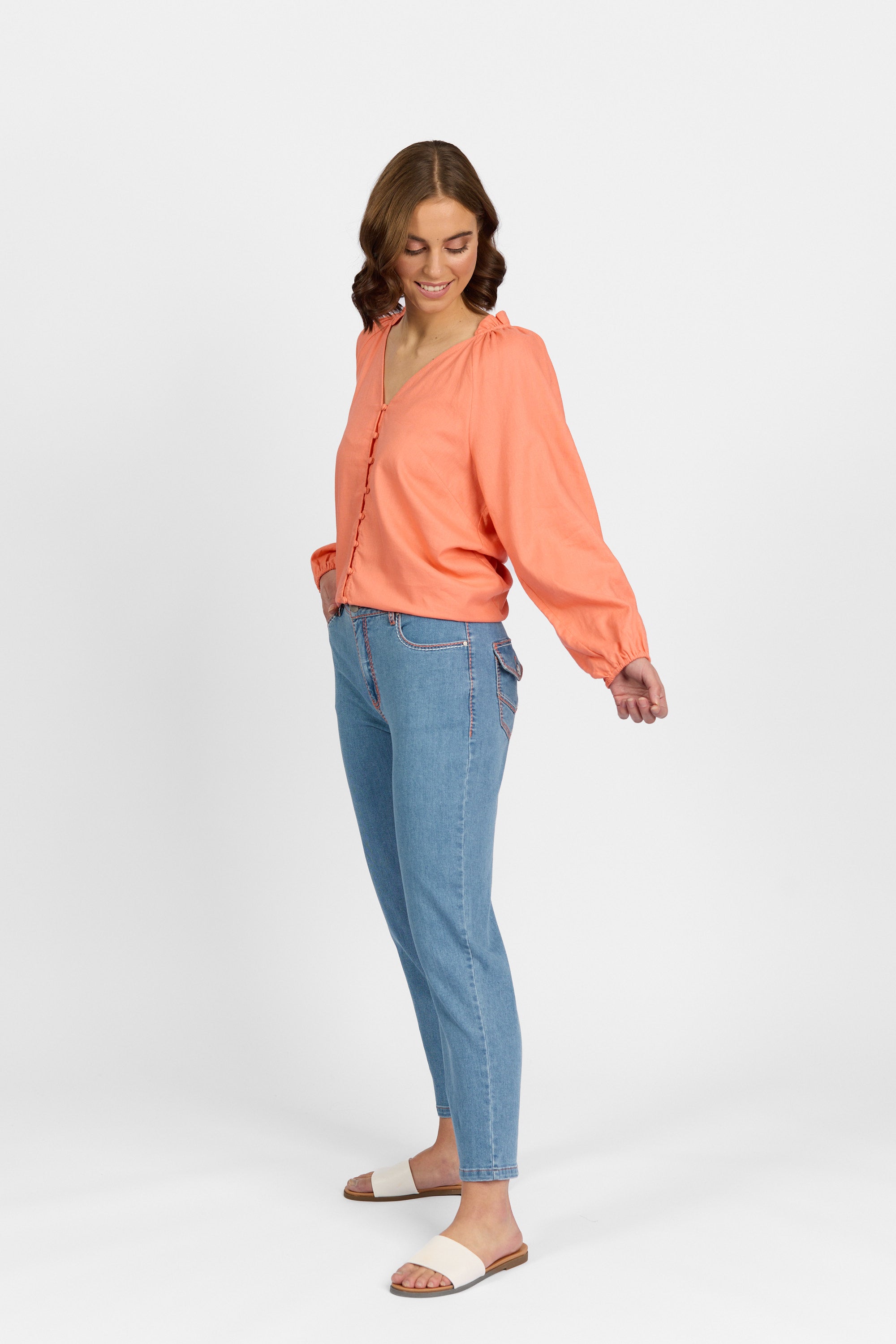 A three-quarter side view of a woman looking down and smiling, wearing palm blue straight-leg ankle-grazer jeans with contrast stitching and flap back pockets, paired with a coral long-sleeve V-neck blouse and white slide sandals.