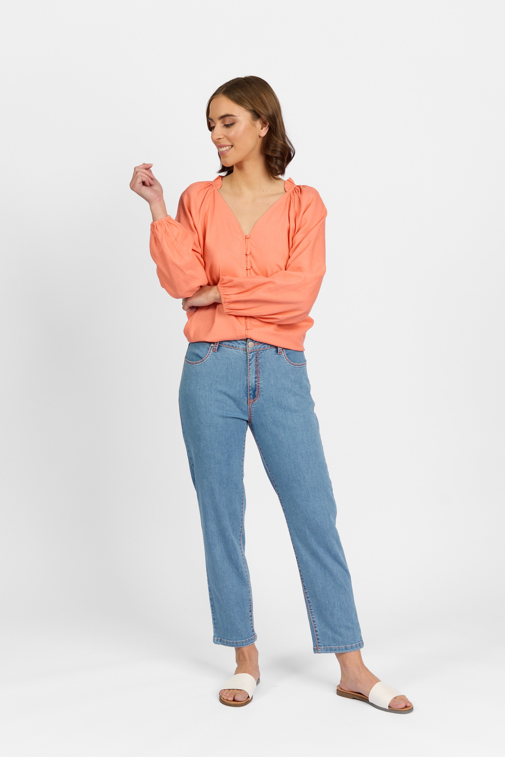 A woman with brown hair smiles looking down with one hand raised, wearing palm blue straight-leg ankle-grazer jeans with contrast stitching, styled with a coral long-sleeve V-neck blouse with ruffled collar and white slide sandals against a light background.