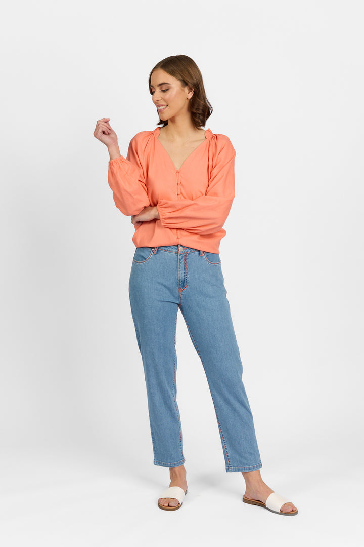 A woman with brown hair smiles looking down with one hand raised, wearing palm blue straight-leg ankle-grazer jeans with contrast stitching, styled with a coral long-sleeve V-neck blouse with ruffled collar and white slide sandals against a light background.