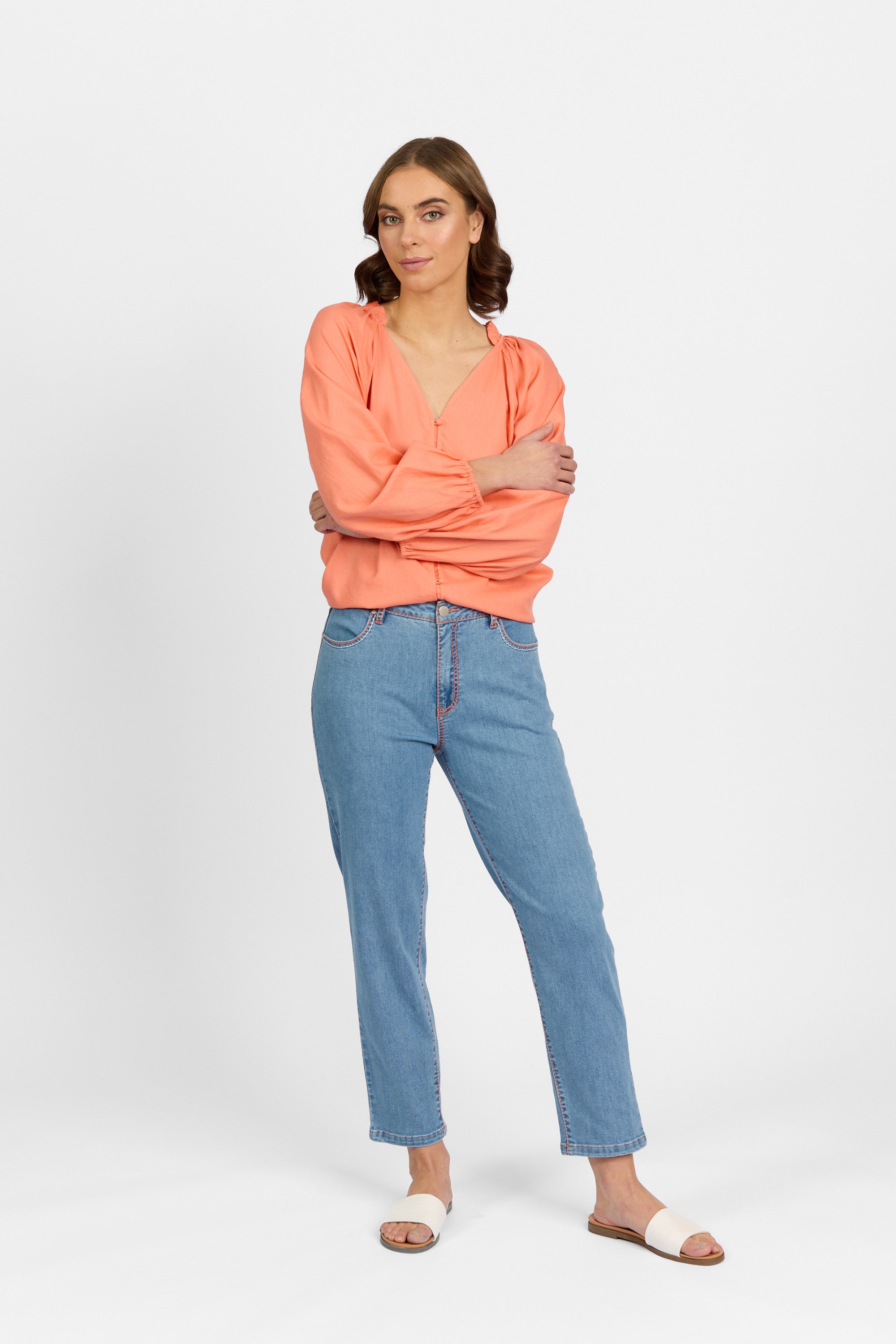 A woman with brown hair stands with arms crossed, wearing palm blue straight-leg ankle-grazer jeans with contrast stitching, paired with a coral long-sleeve V-neck blouse and white slide sandals against a plain light background.