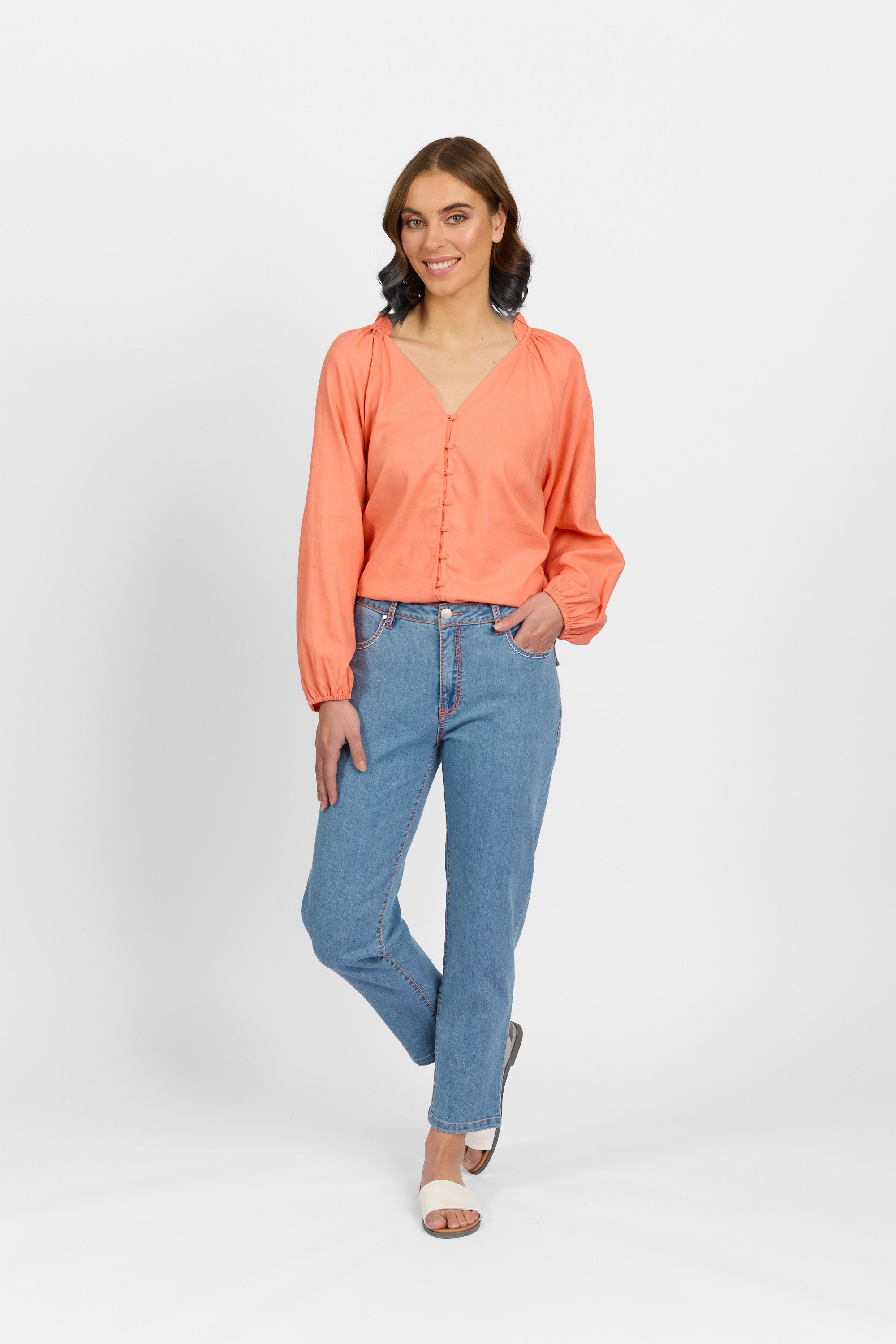 A woman with brown wavy hair smiles with one hand in her pocket, wearing palm blue straight-leg ankle-grazer jeans, styled with a coral long-sleeve ruffle-neck blouse and white heeled sandals against a plain light background.