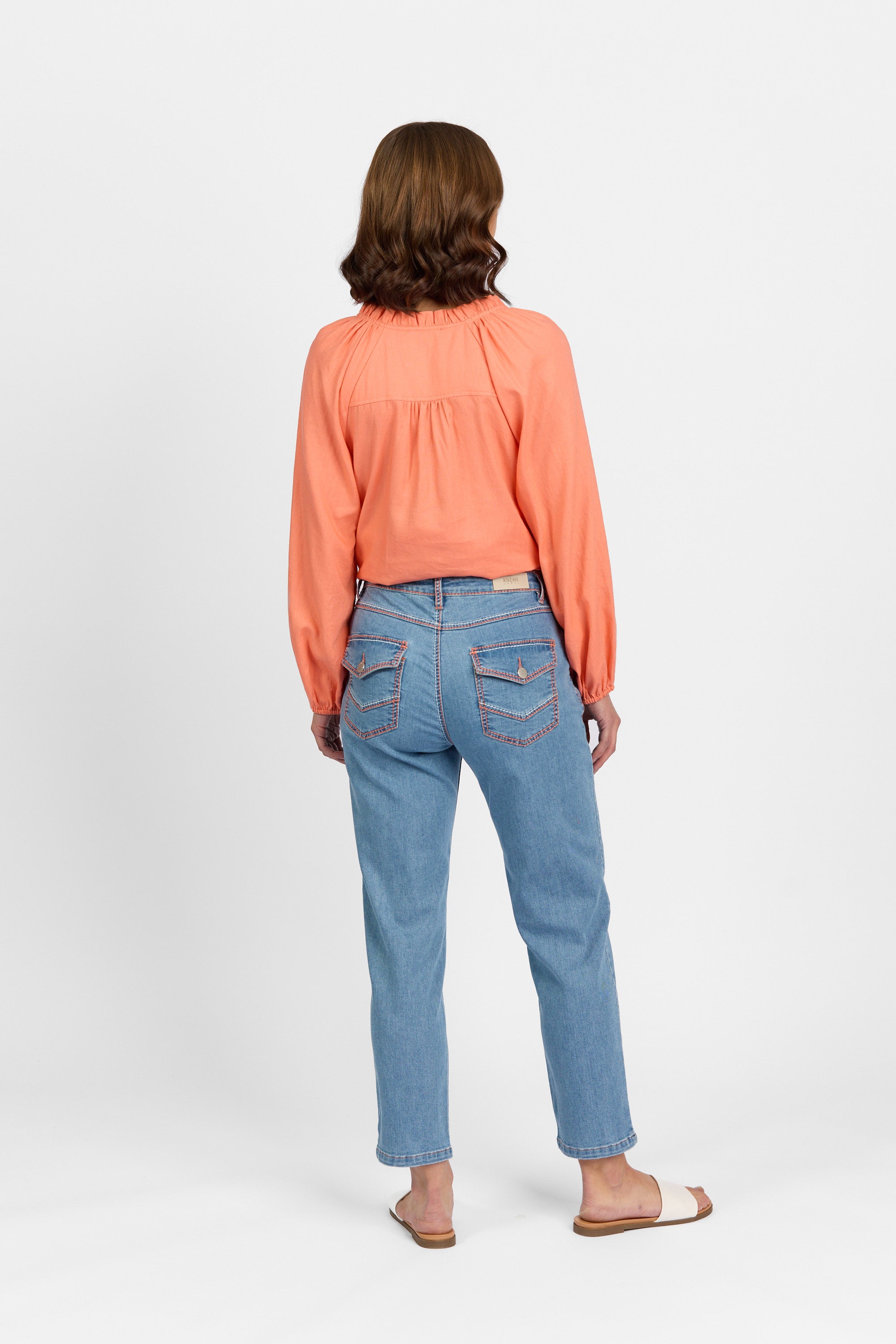 Back view of a woman with brown wavy hair wearing palm blue straight-leg ankle-grazer jeans with contrast stitching, flap back pockets and a leather KNEWE brand patch at the waistband, paired with a coral long-sleeve blouse and white flat sandals.