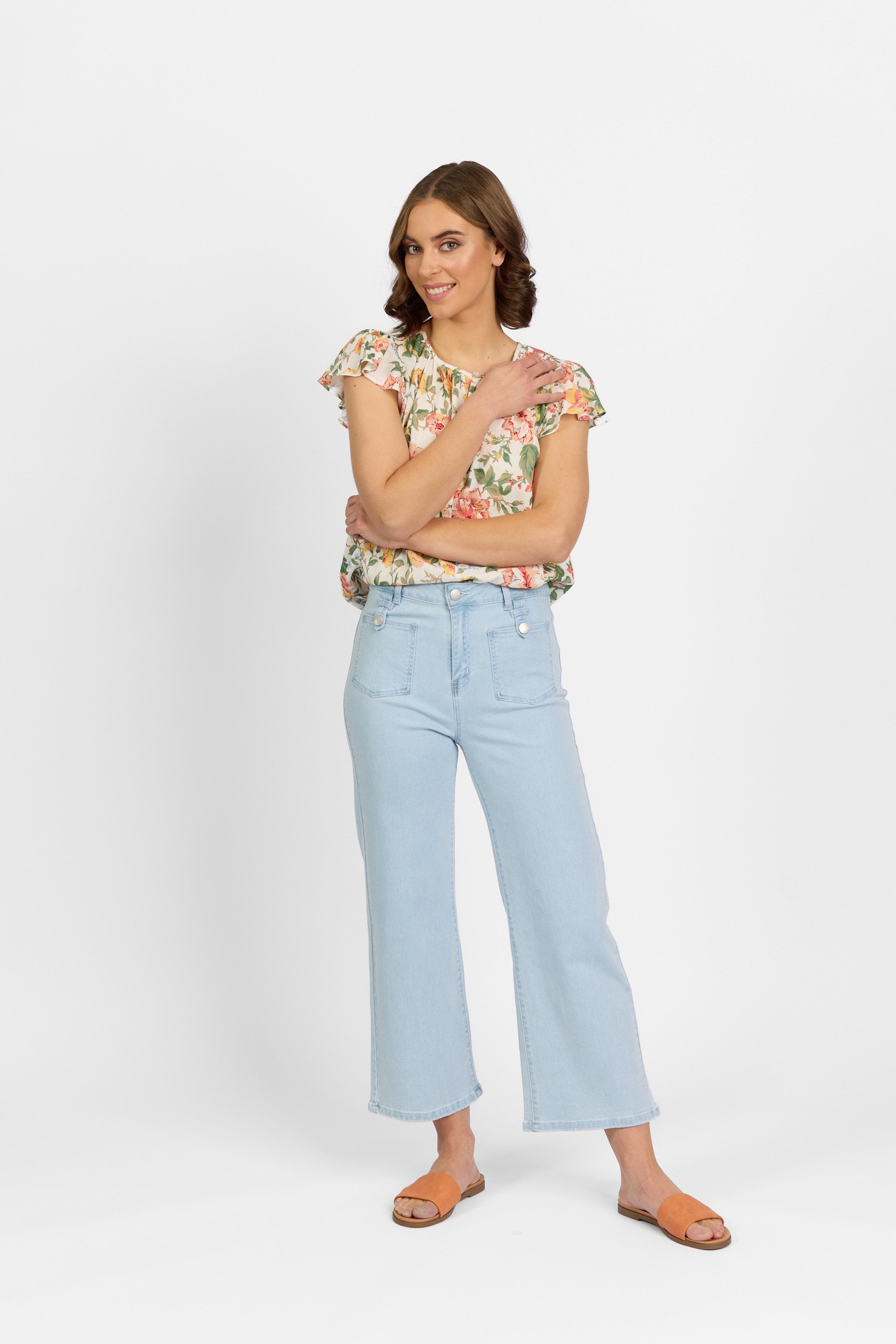 A woman with brown hair smiles with her arms crossed, wearing light wash wide-leg cropped jeans with front patch pockets and metal button tab detailing, paired with a floral ruffle blouse and tan slide sandals against a plain light background.