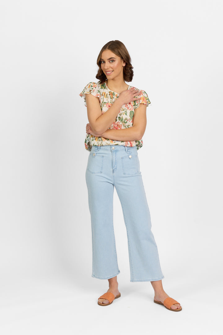 A woman with brown hair smiles with her arms crossed, wearing light wash wide-leg cropped jeans with front patch pockets and metal button tab detailing, paired with a floral ruffle blouse and tan slide sandals against a plain light background.