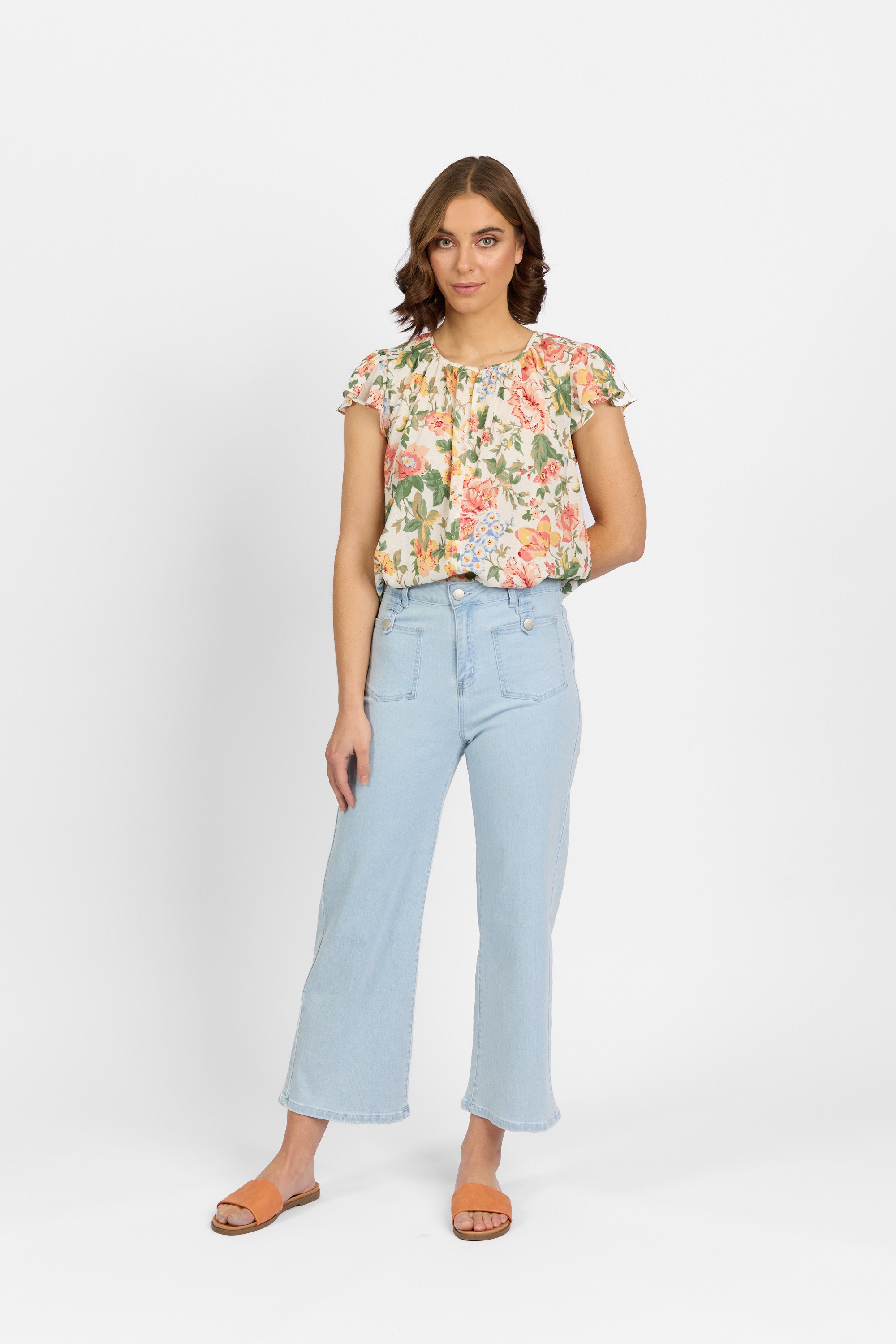 A woman with brown hair stands facing forward wearing light wash wide-leg cropped jeans with front patch pockets and metal hardware, paired with a floral short-sleeve top and tan slide sandals against a plain light background.