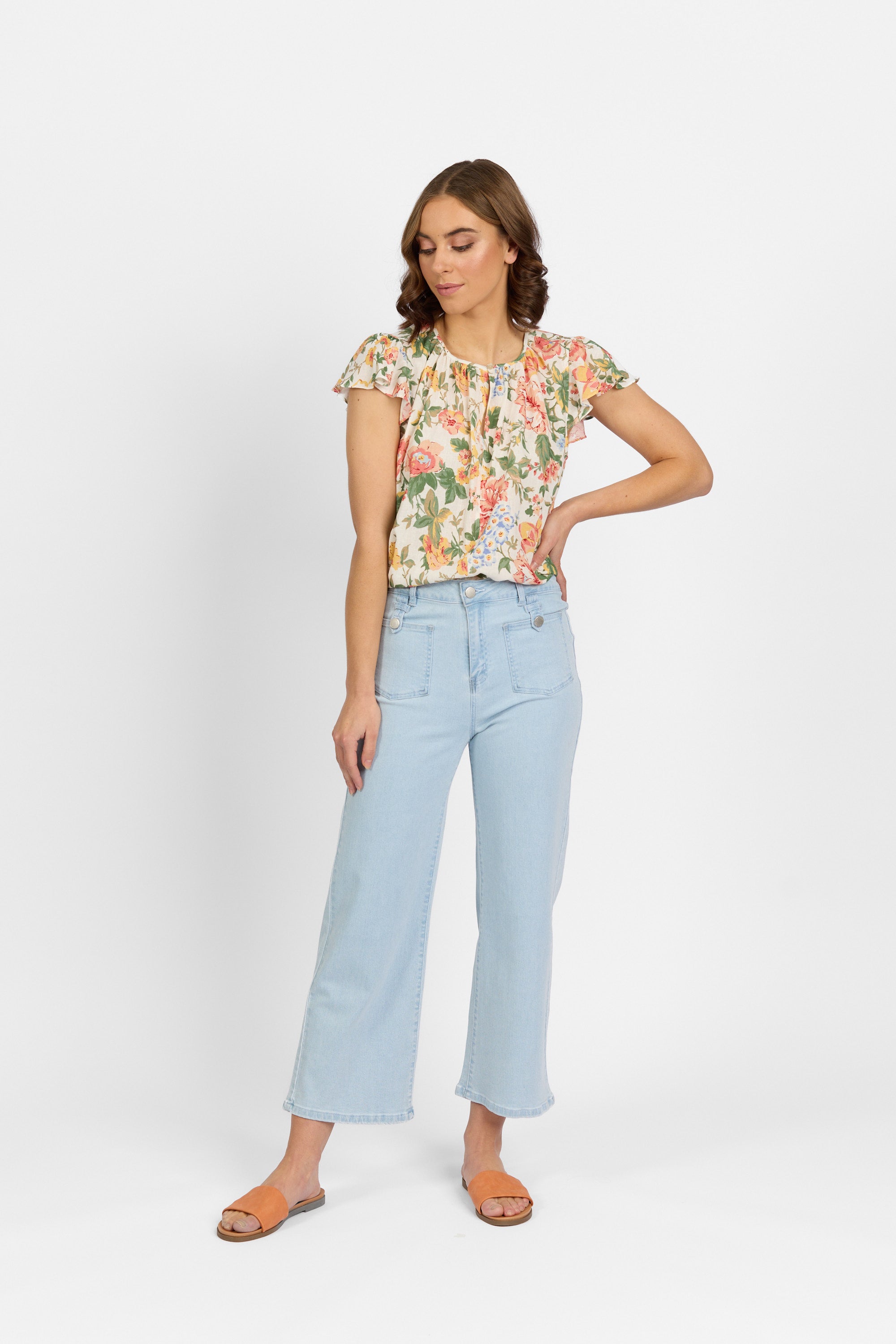 A woman with brown hair looks down with one hand on her hip, wearing light wash wide-leg cropped jeans with front patch pockets and metal button tabs, styled with a floral ruffle-sleeve top and tan slide sandals against a light background.