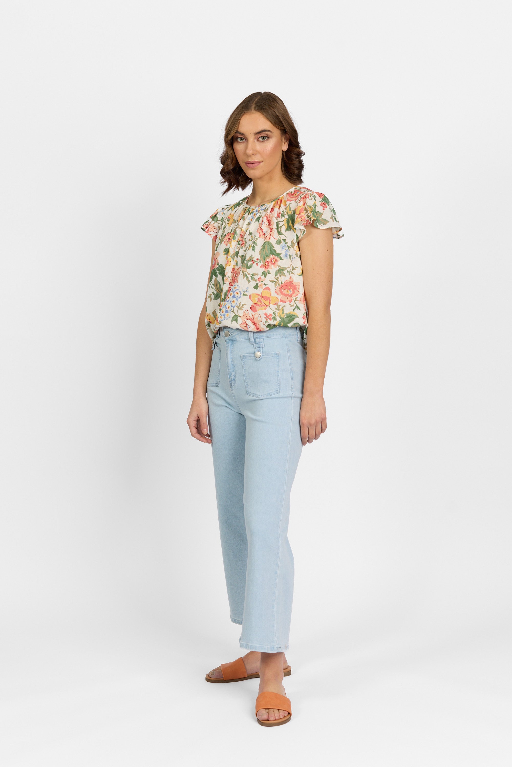 A woman with brown hair stands facing slightly to the side wearing light wash wide-leg cropped jeans with prominent front patch pockets, paired with a floral short-sleeve ruffle blouse and tan slide sandals against a plain light background.