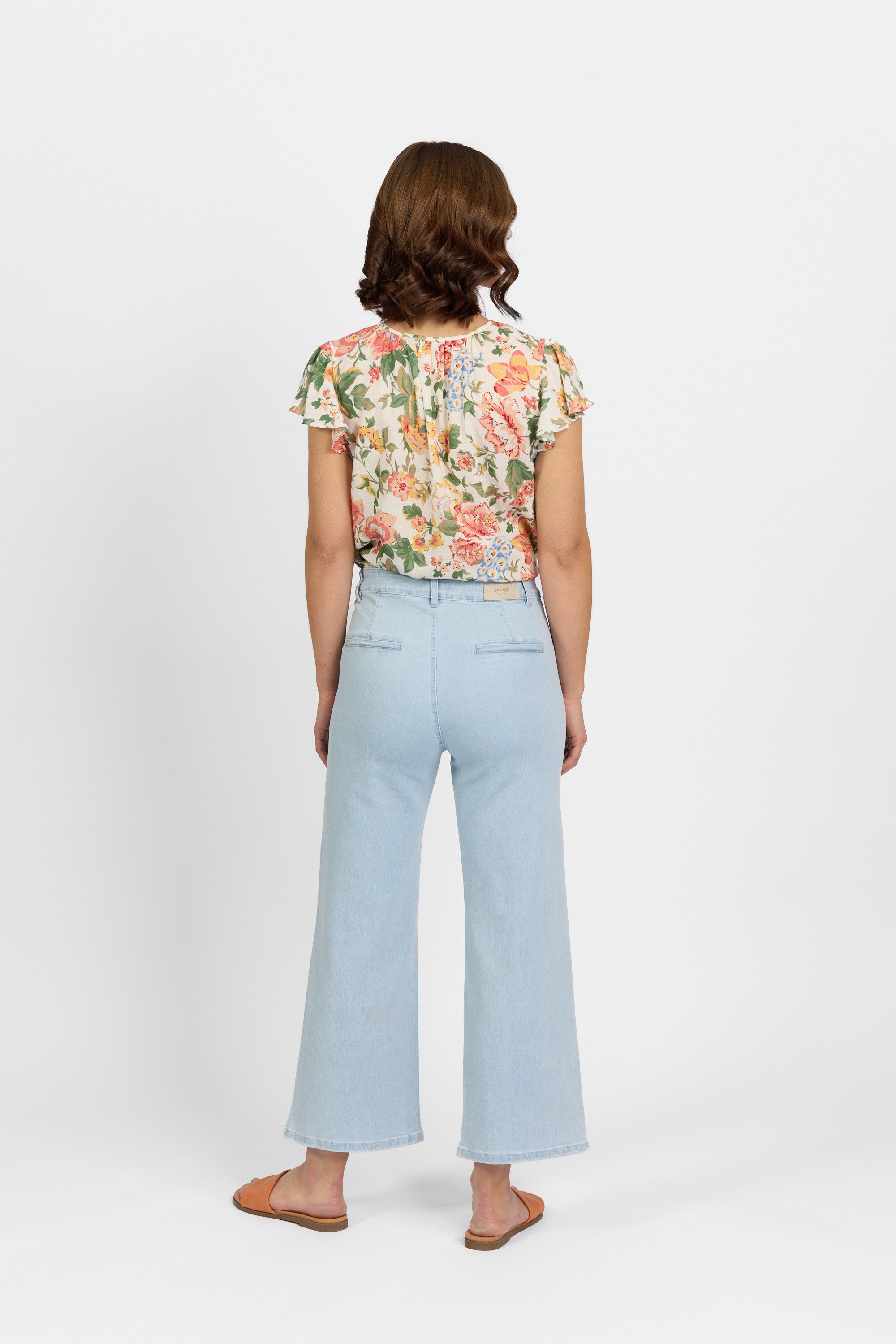 Back view of a woman wearing light wash wide-leg cropped jeans with faux welt back pockets and a leather KNEWE brand patch on the waistband, paired with a floral short-sleeve top and tan slide sandals against a light background.