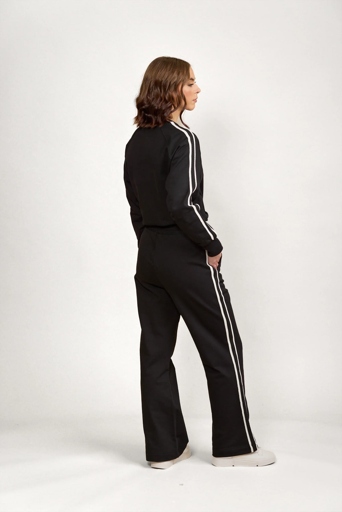 Woman wearing black wide leg pants and track jacket with white side stripes, side view