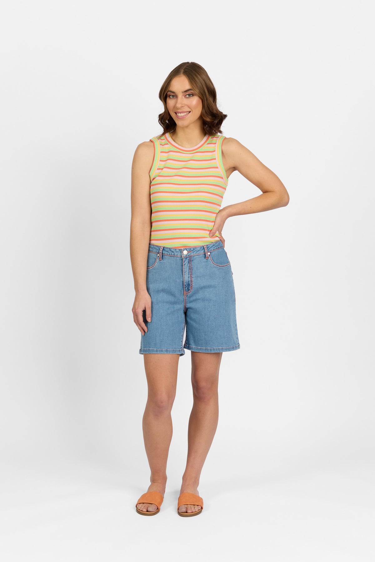 A woman with brown wavy hair smiles at the camera wearing palm blue denim shorts with contrast stitching, paired with a multicoloured striped ribbed tank top and tan slide sandals against a plain light background.