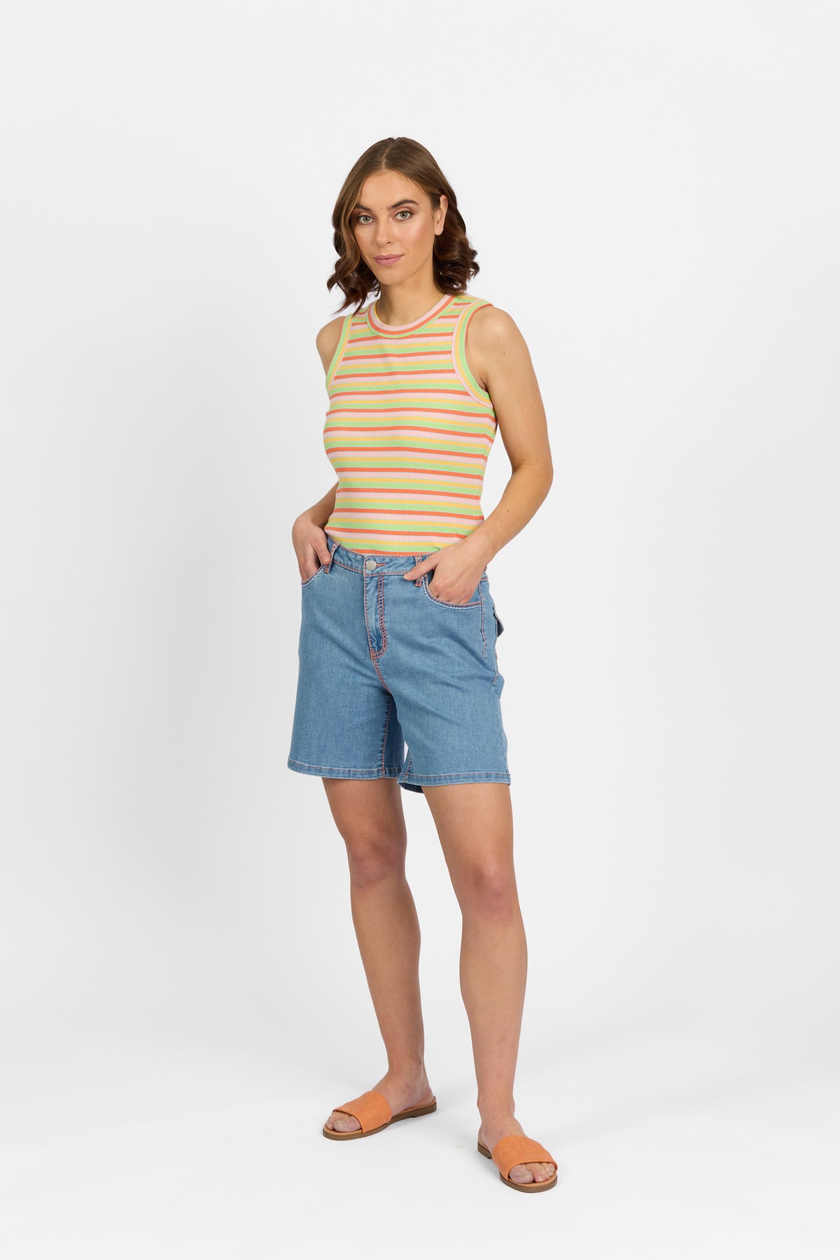 A woman with brown wavy hair stands with both hands in the pockets of palm blue denim shorts with contrast stitching, paired with a multicoloured striped tank top and tan slide sandals against a plain light background.